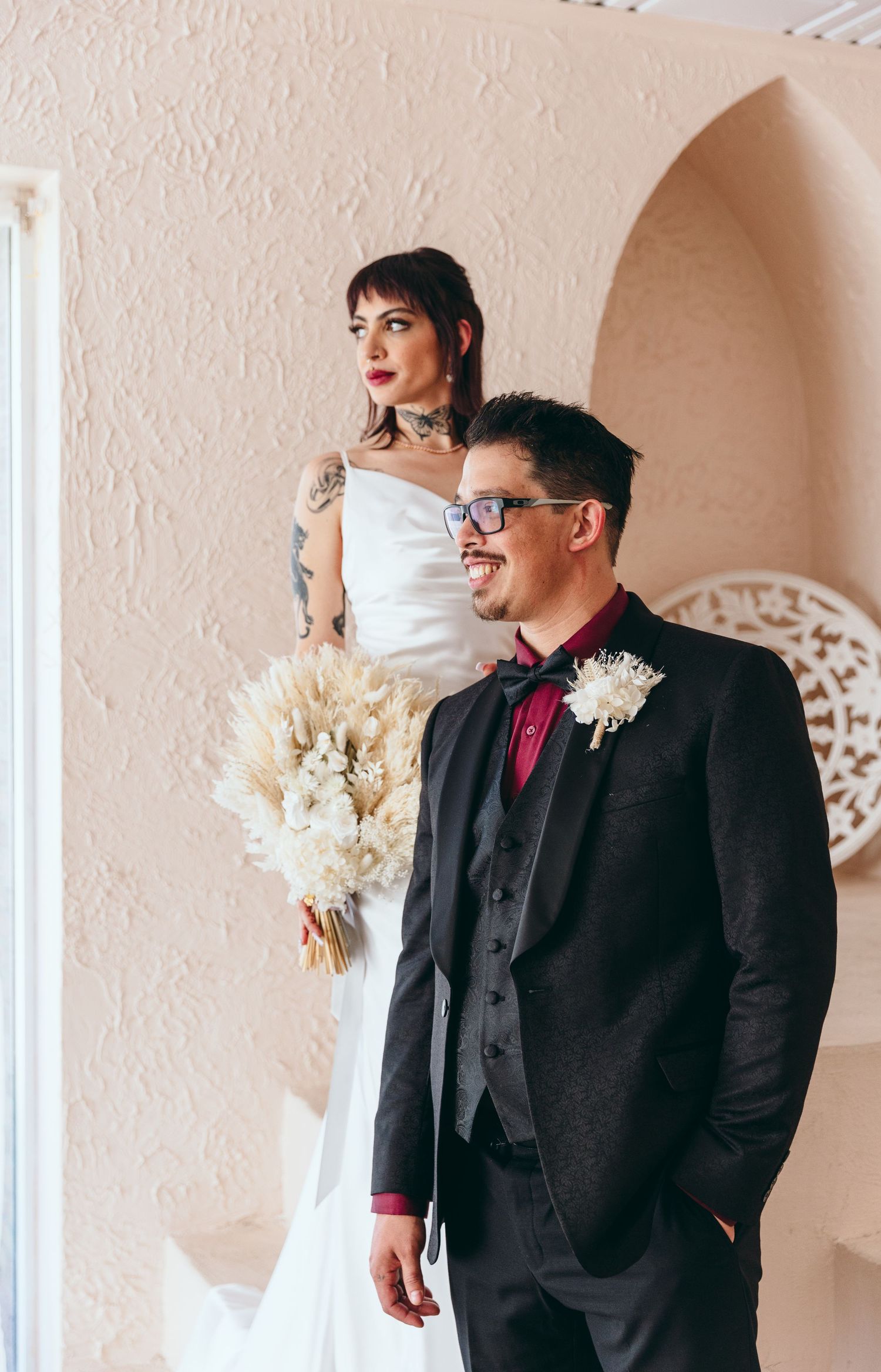 Wedding couple poses in front of arched windows with white decor and natural light.