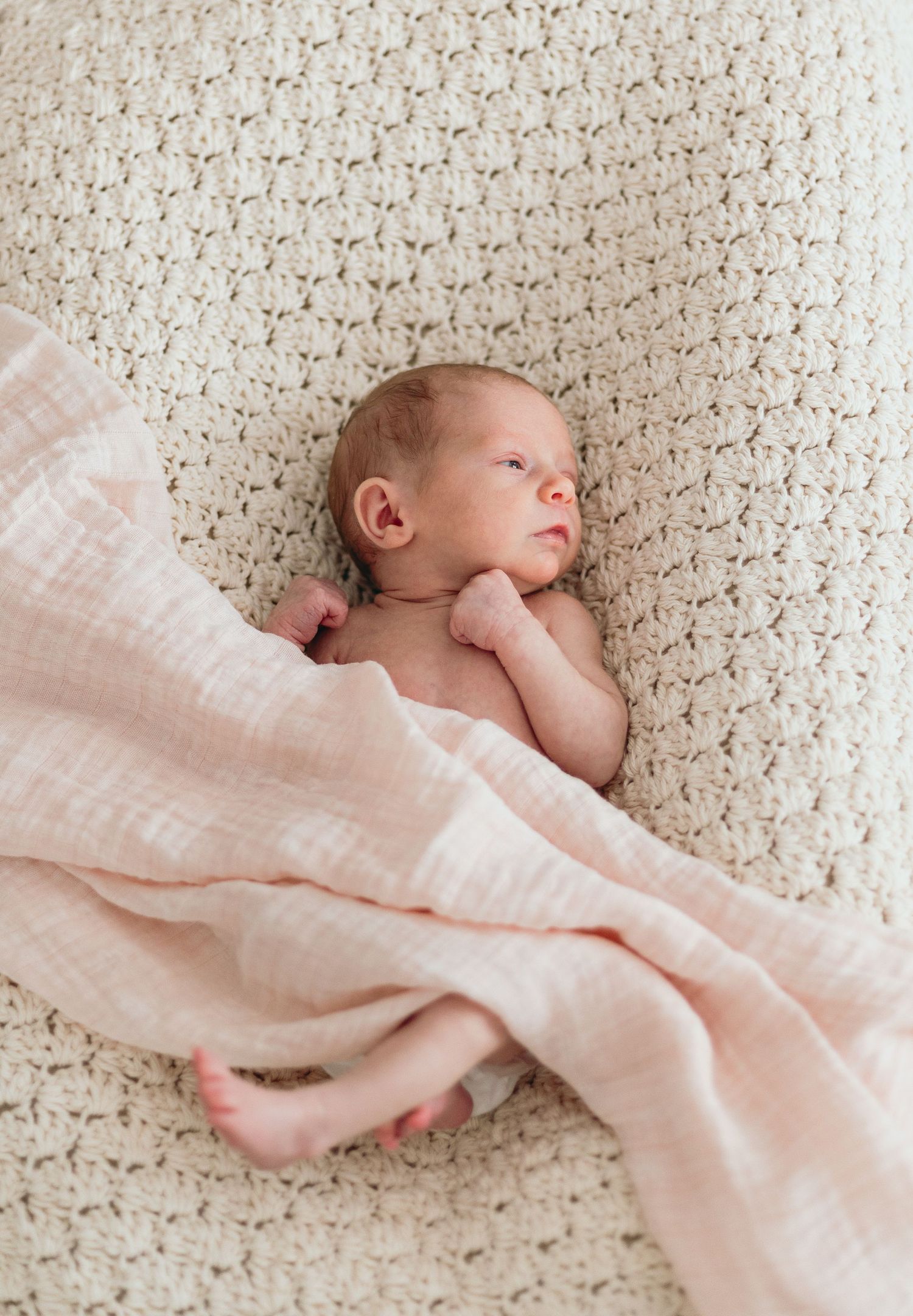 Baby lying on textured cream blanket with soft pink muslin wrap in natural light.