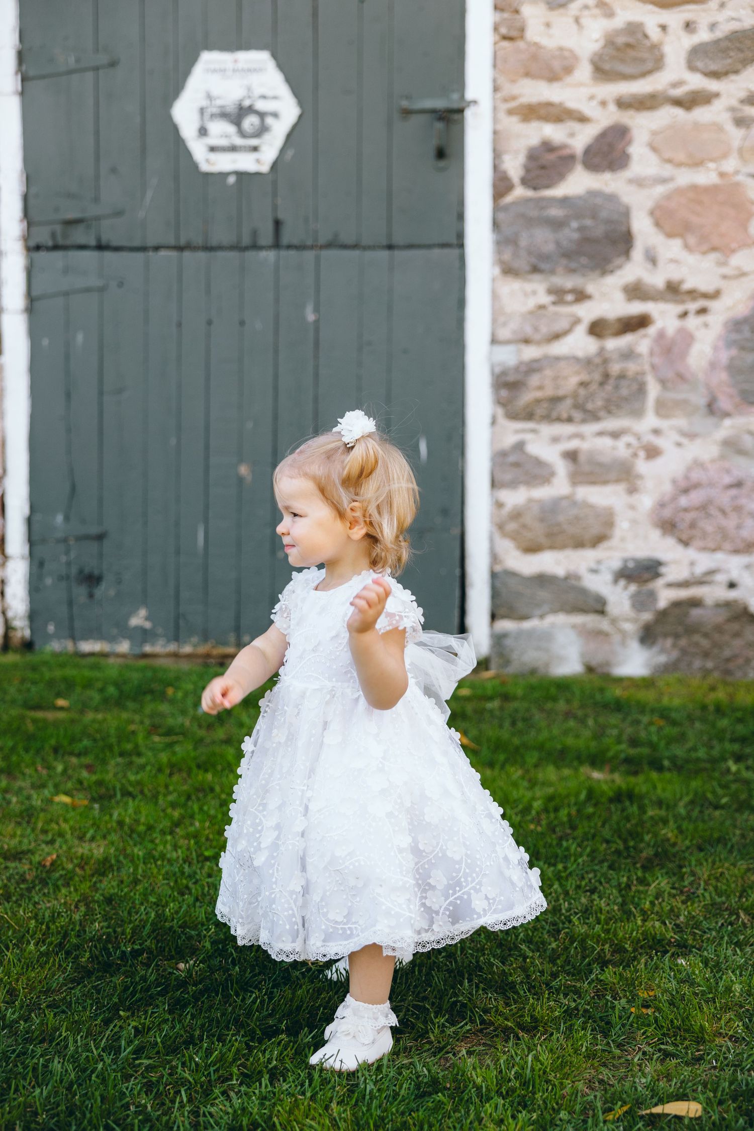 A small child in a white lace dress twirls on grass near a stone building with green doors.