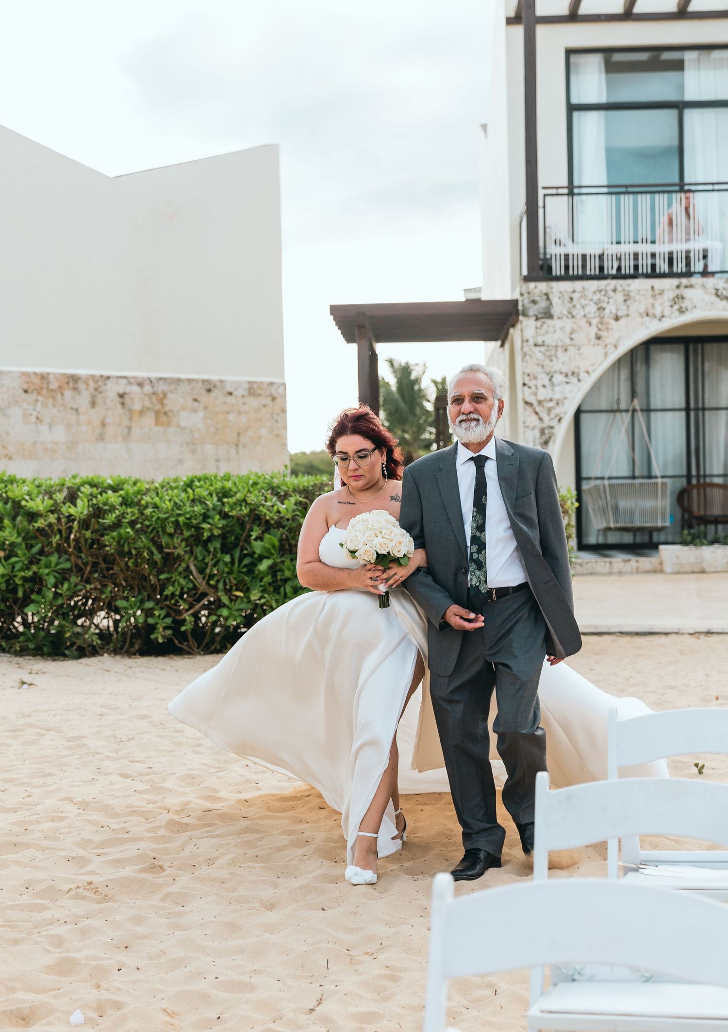 Wedding party members pose on sandy beach with resort building in background.