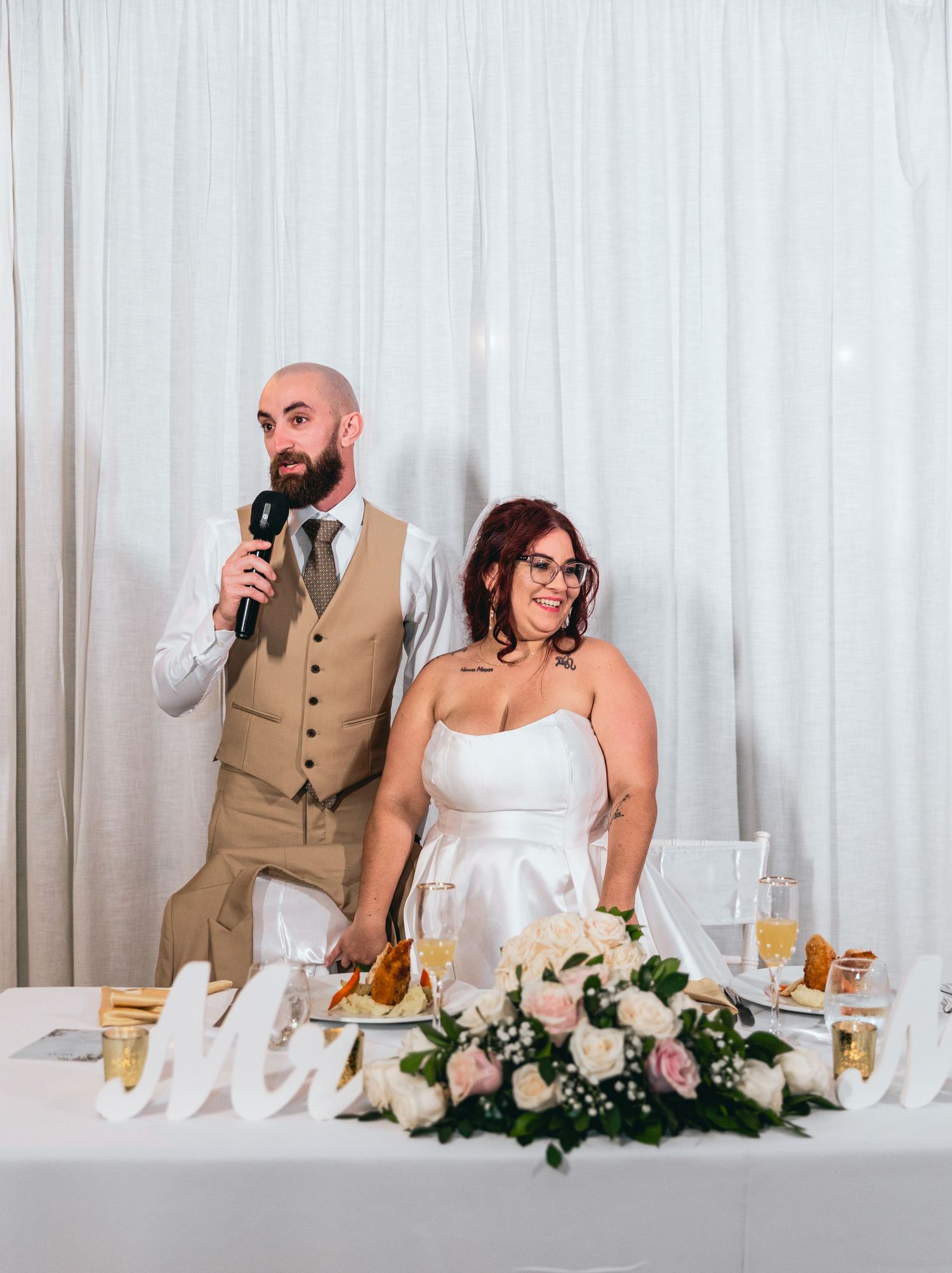 A couple stands at their wedding reception head table giving speeches.