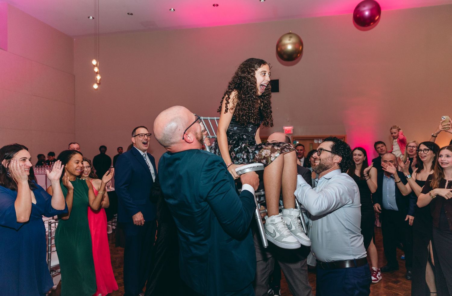 Wedding reception dance floor filled with guests celebrating under pink lighting and disco balls.