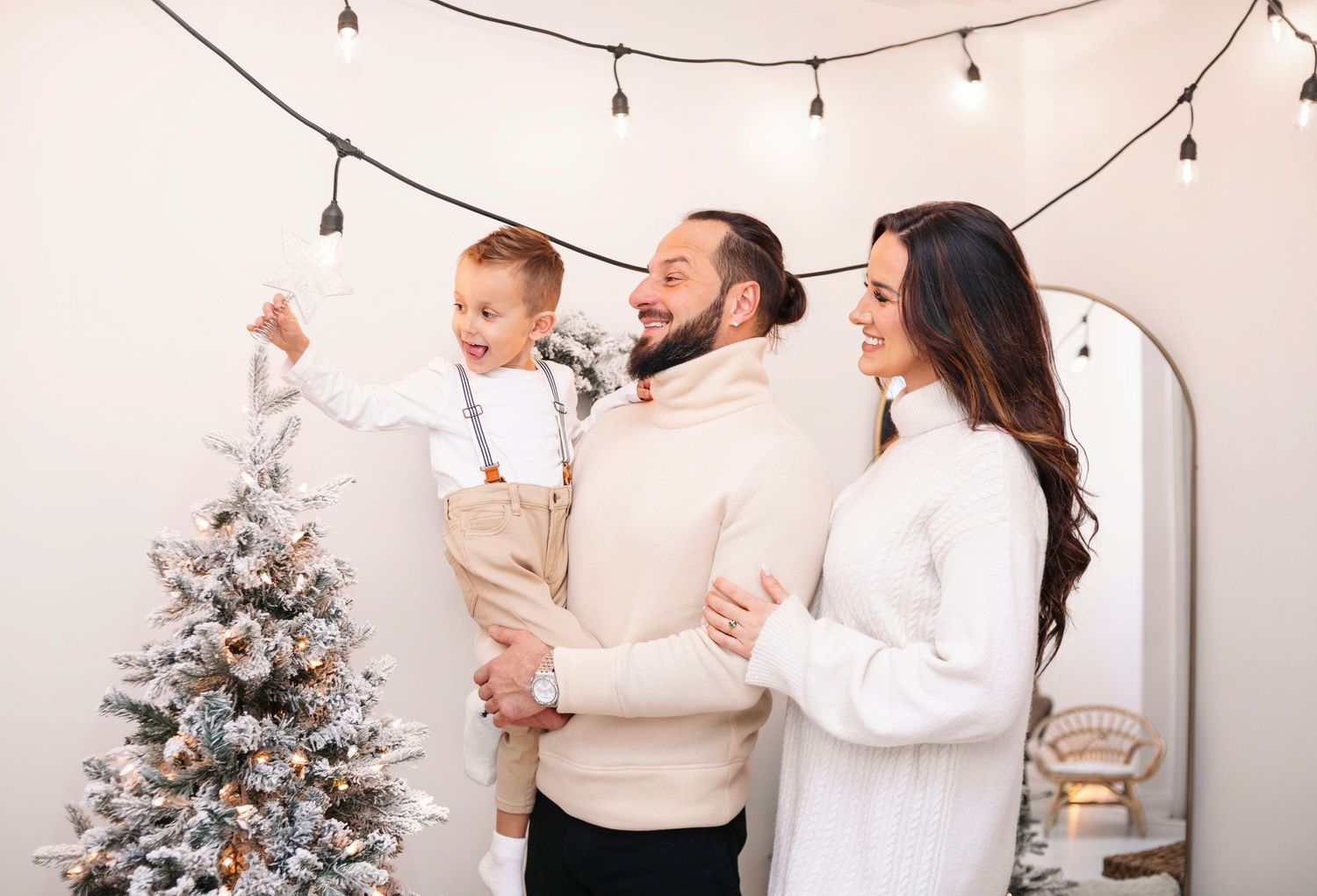 Family decorating a flocked Christmas tree with string lights, father holding toddler son, mother smiling nearby.