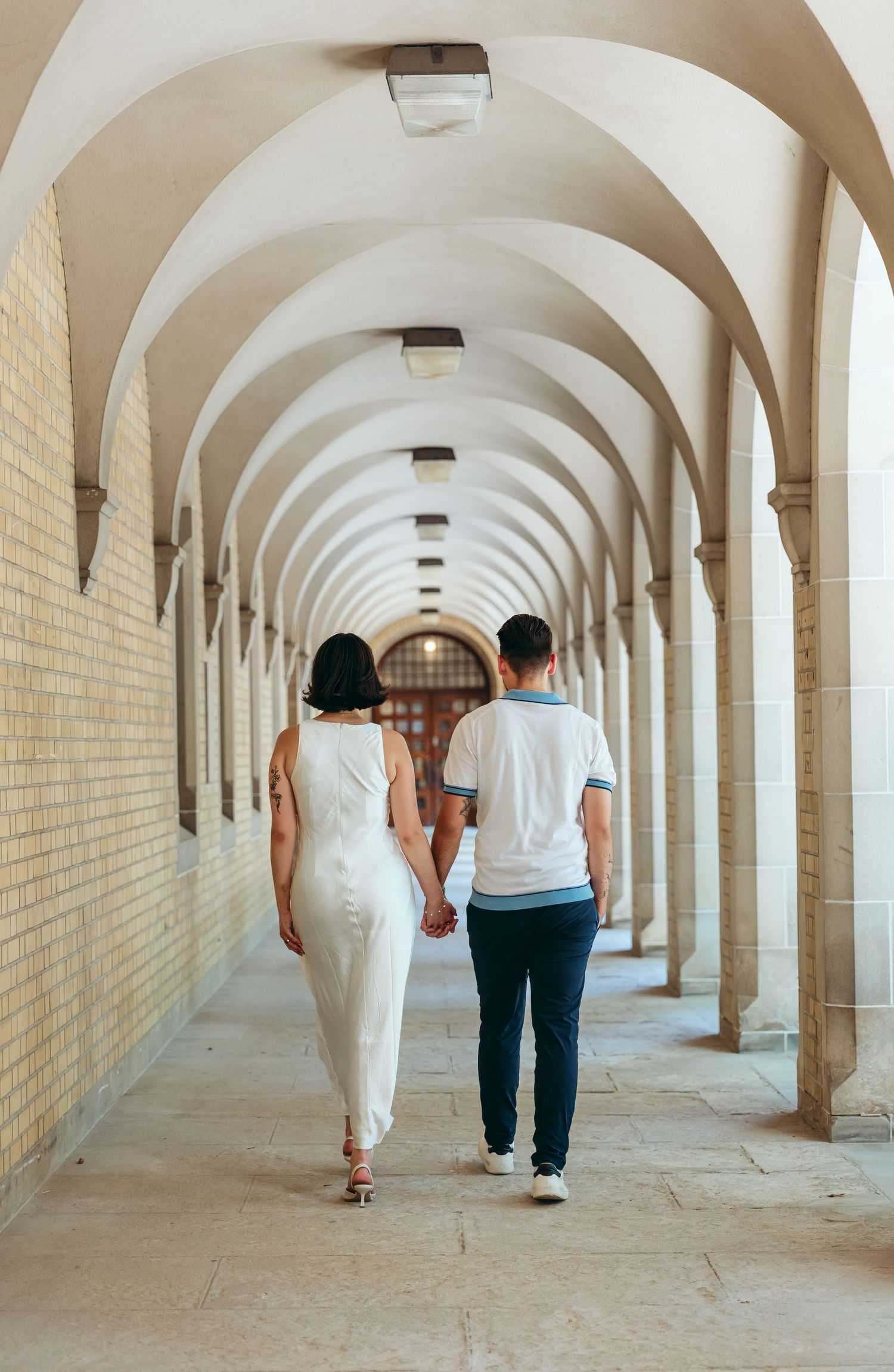 Couple strolling down long arched corridor with white walls and columns.