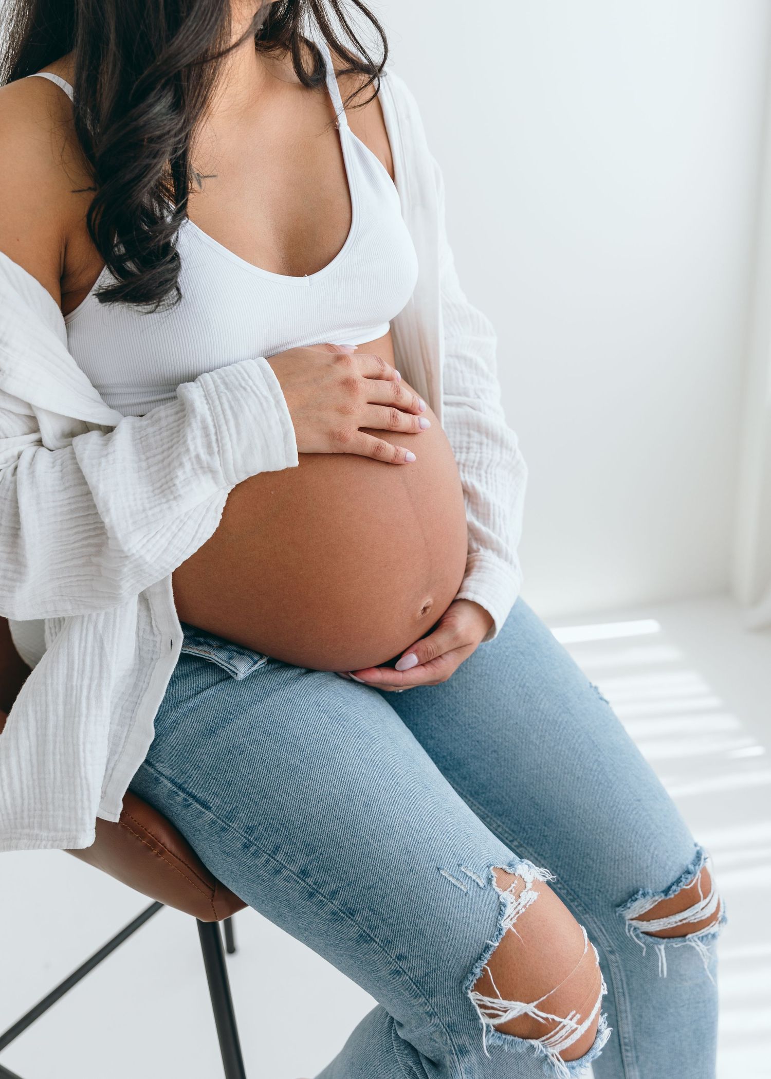 Maternity photoshoot showing pregnant woman in white top and ripped jeans against white background.