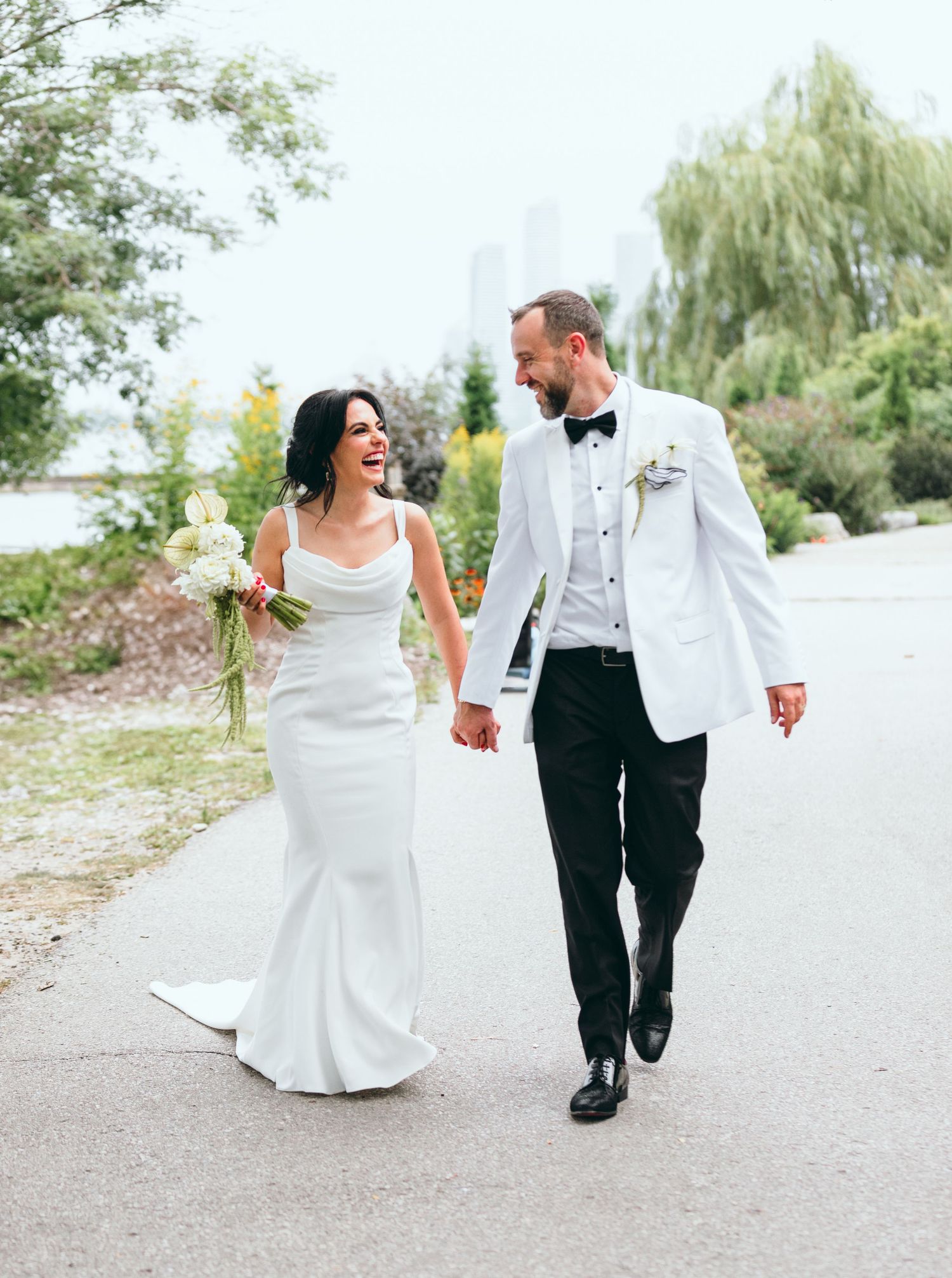 A wedding couple walks hand in hand along a sandy beach path in elegant attire.