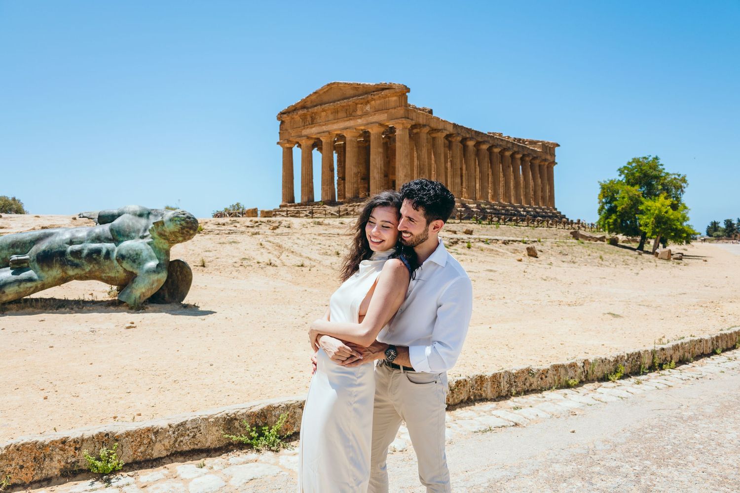 A couple embraces at the Temple of Concordia in Agrigento, Sicily with ancient Greek architecture behind them.