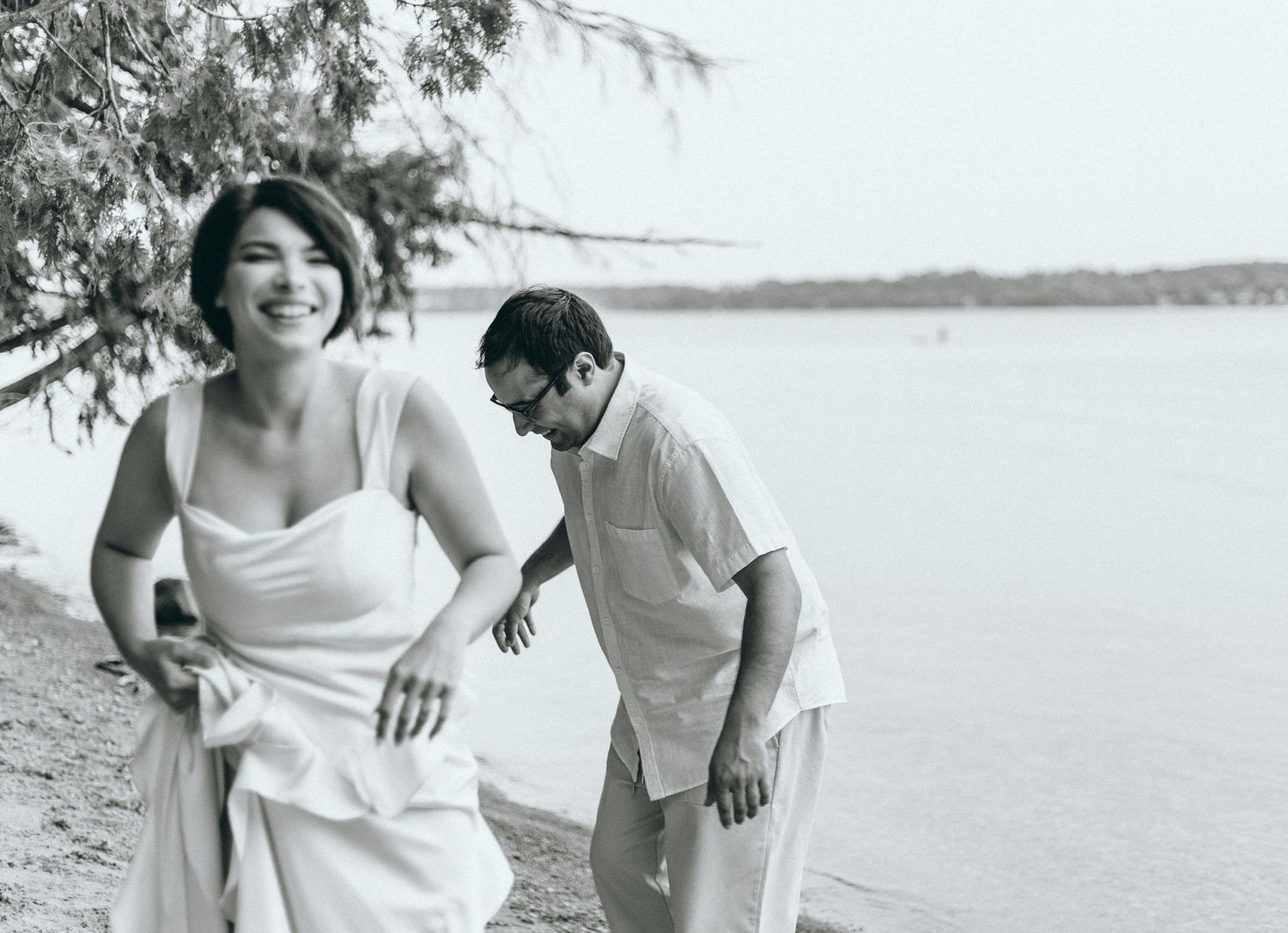 Couple sharing a joyful moment on a beach with water and trees in background.