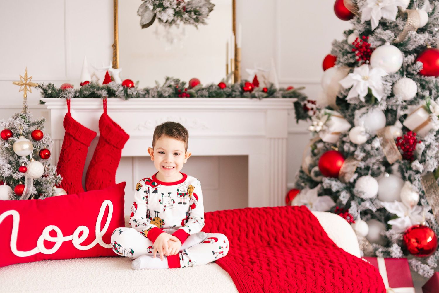 Young boy in Christmas pajamas sits on a red bed near a decorated tree and fireplace with holiday stockings.