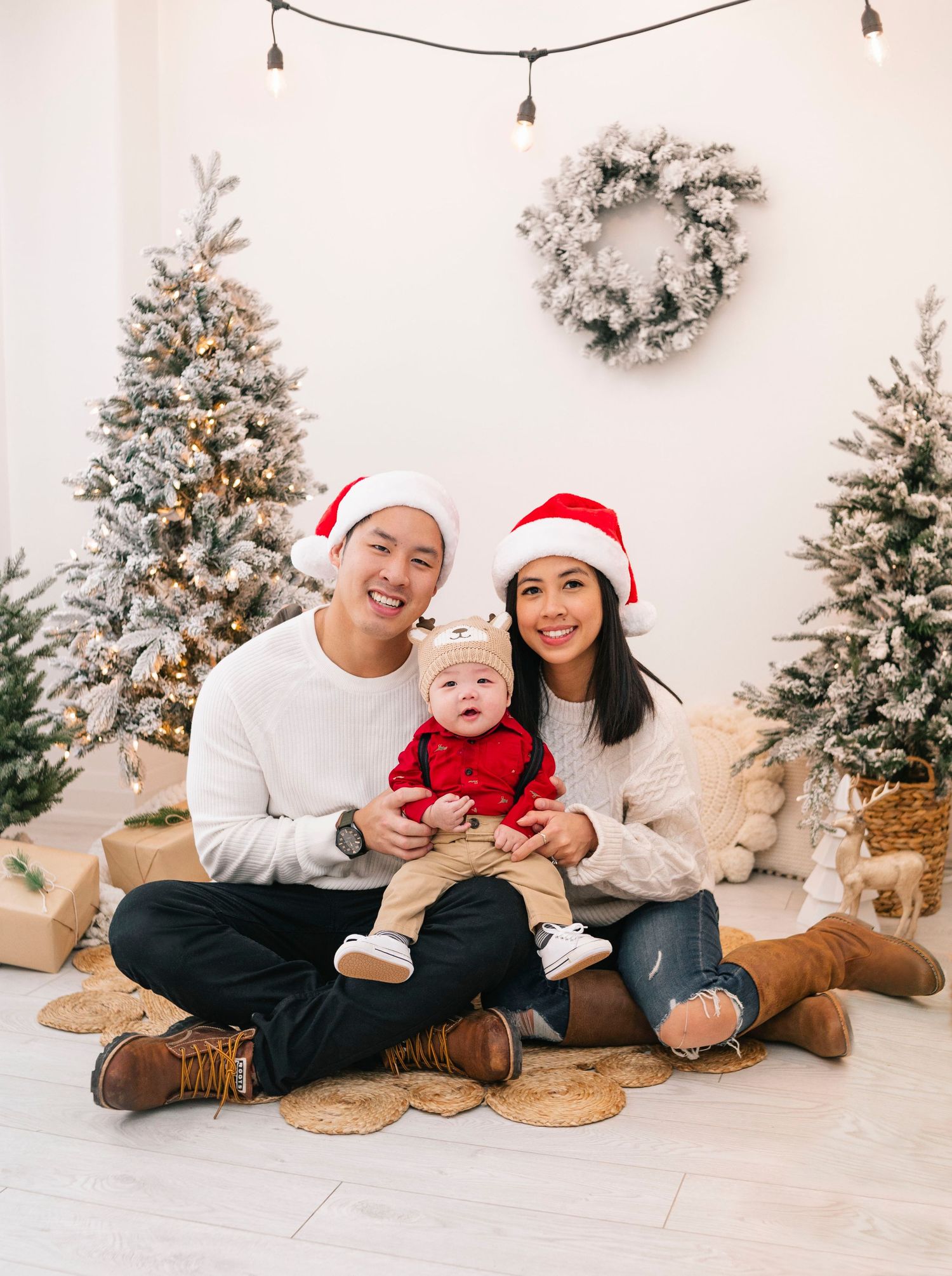 Family in Santa hats poses with baby in red outfit before snowy Christmas trees and wreath backdrop.
