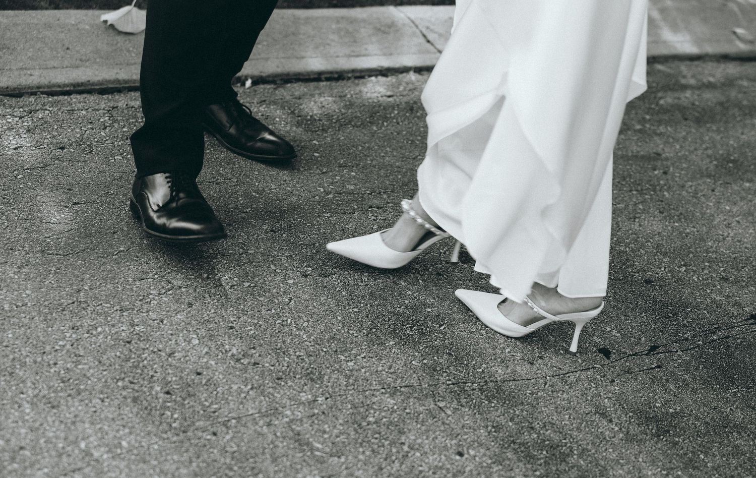 Close-up of formal shoes and dress hems on pavement.