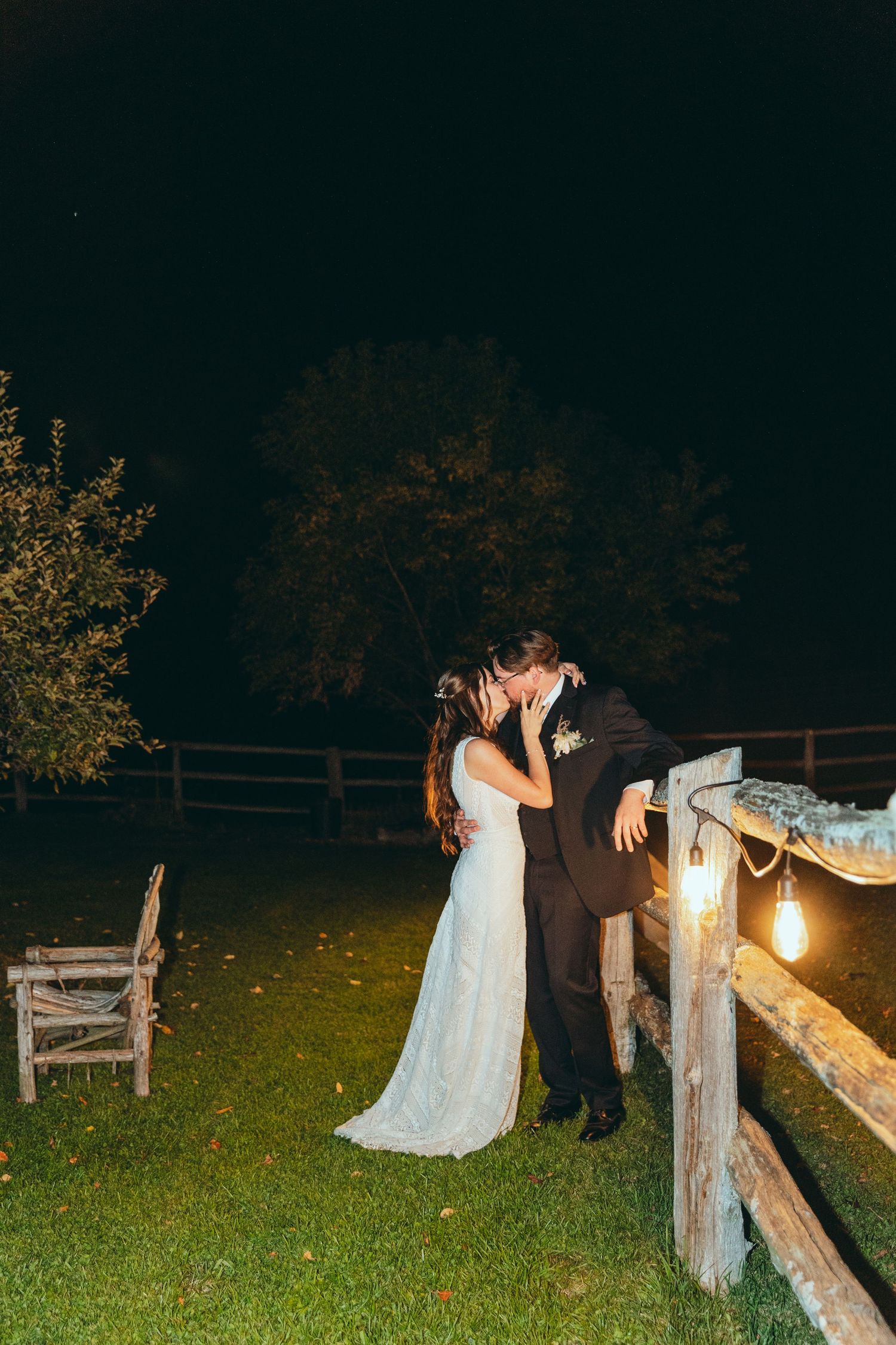 Evening wedding portraits with string lights illuminating couple by wooden fence.