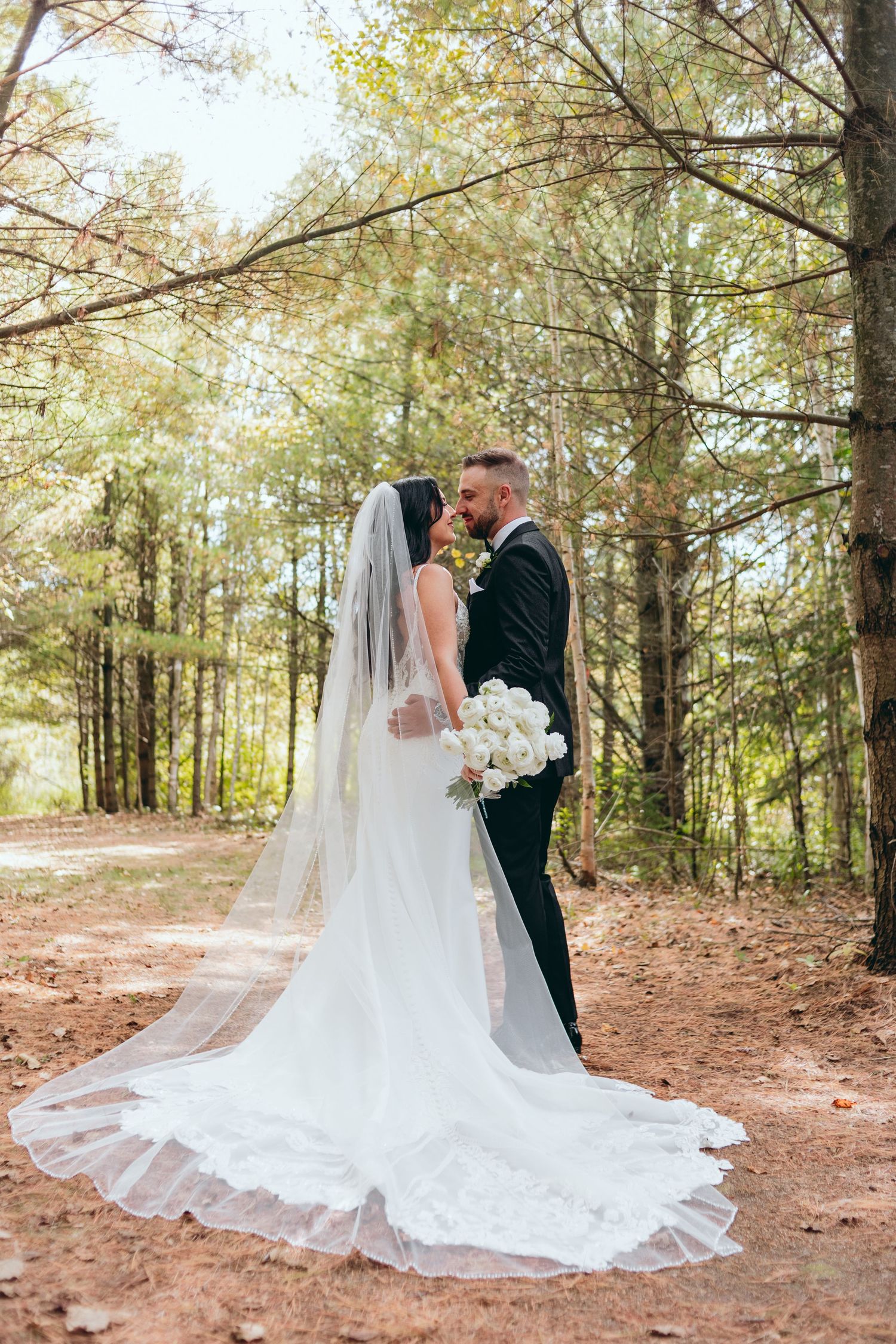 Bride and groom embrace in sunlit forest with flowing cathedral veil trailing behind.