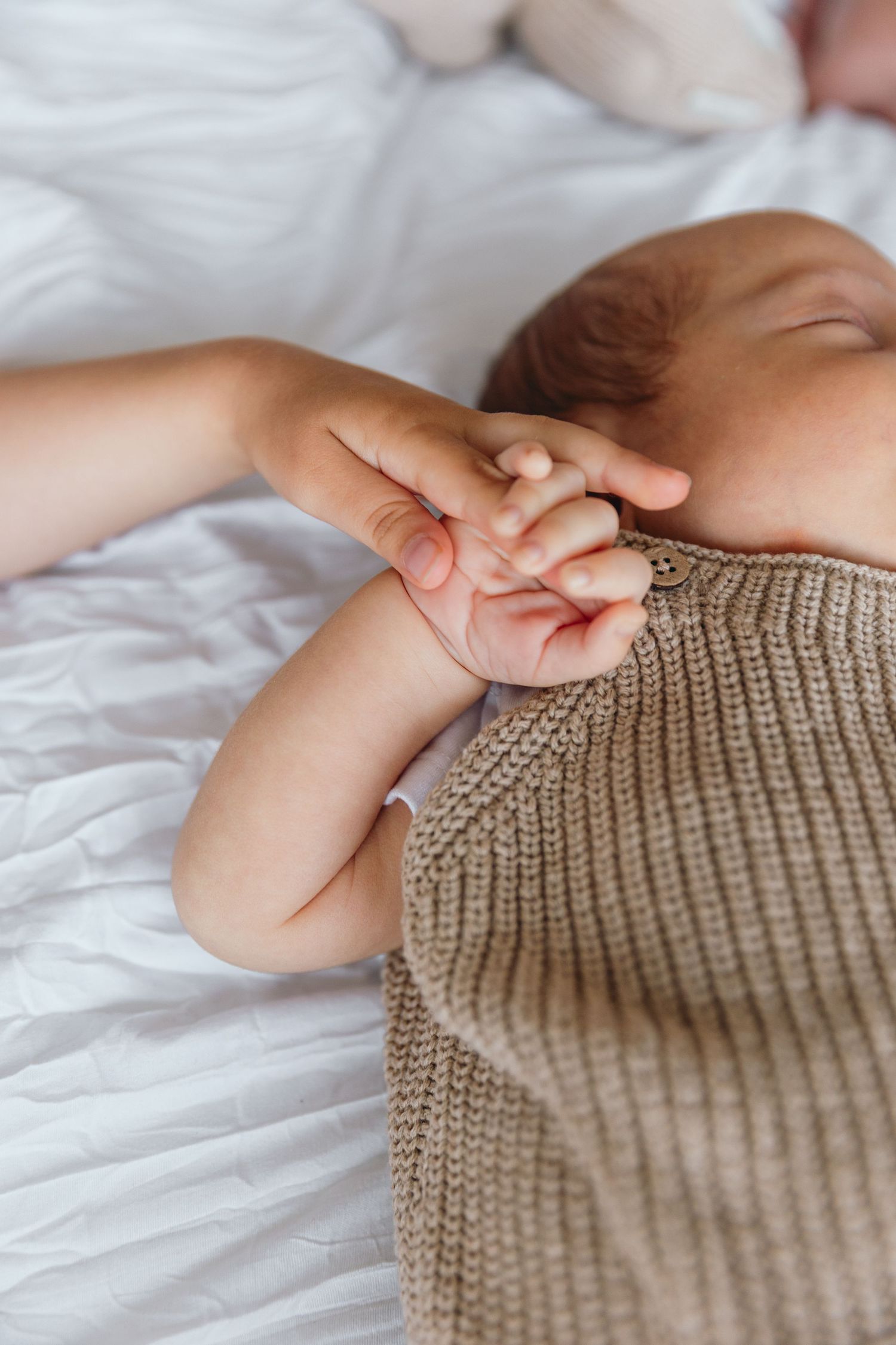 Close up of small hands holding each other on white bedding with knit clothing visible.