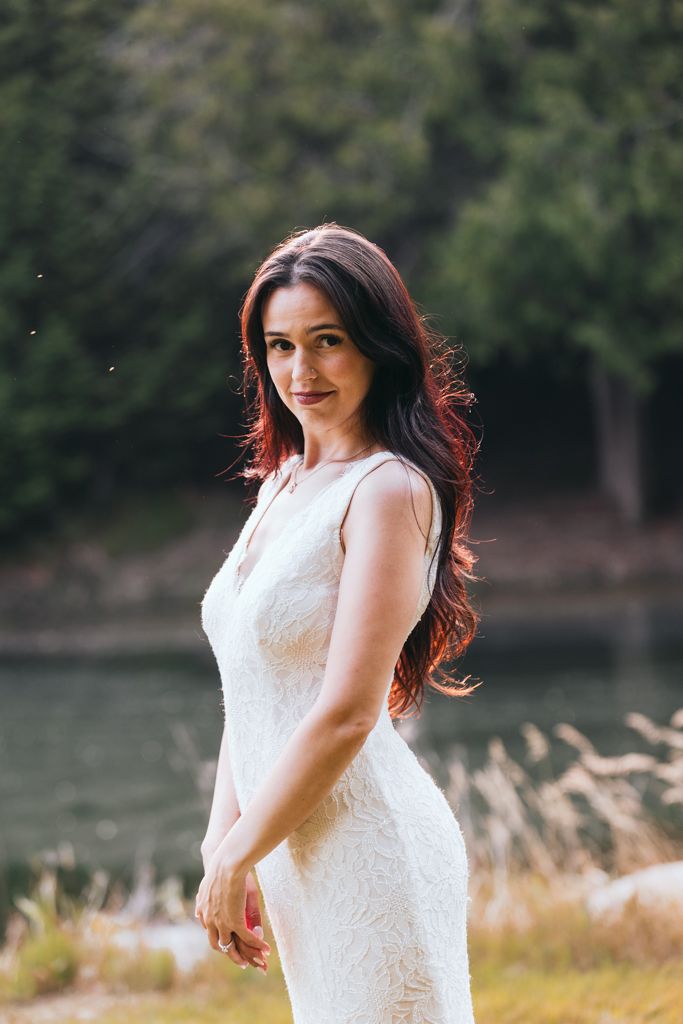 Elegant bride in fitted lace wedding dress poses by lakeside with natural backdrop.