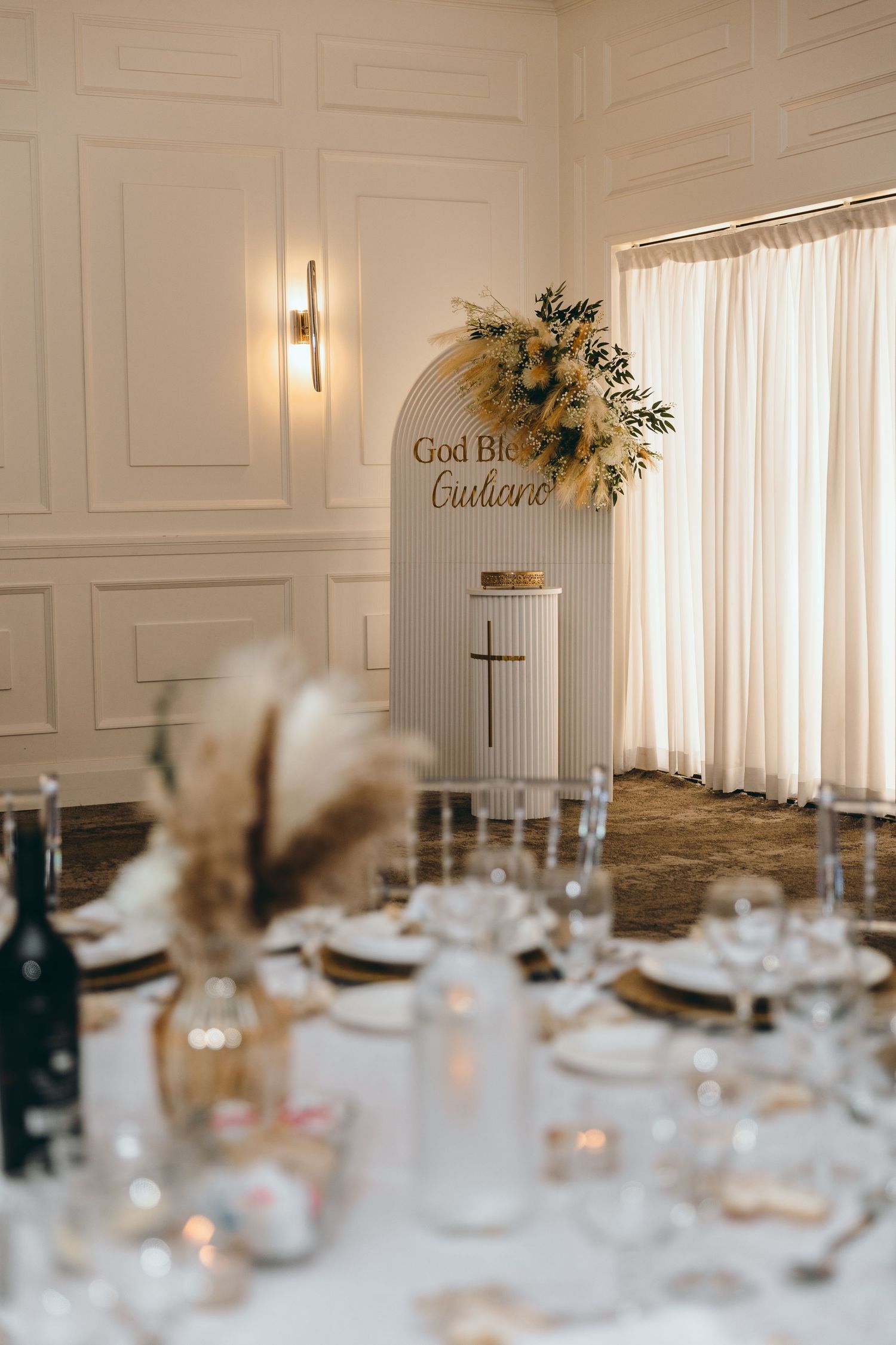 Elegant table setting with pampas grass centerpieces under a white arched backdrop with gold lettering.