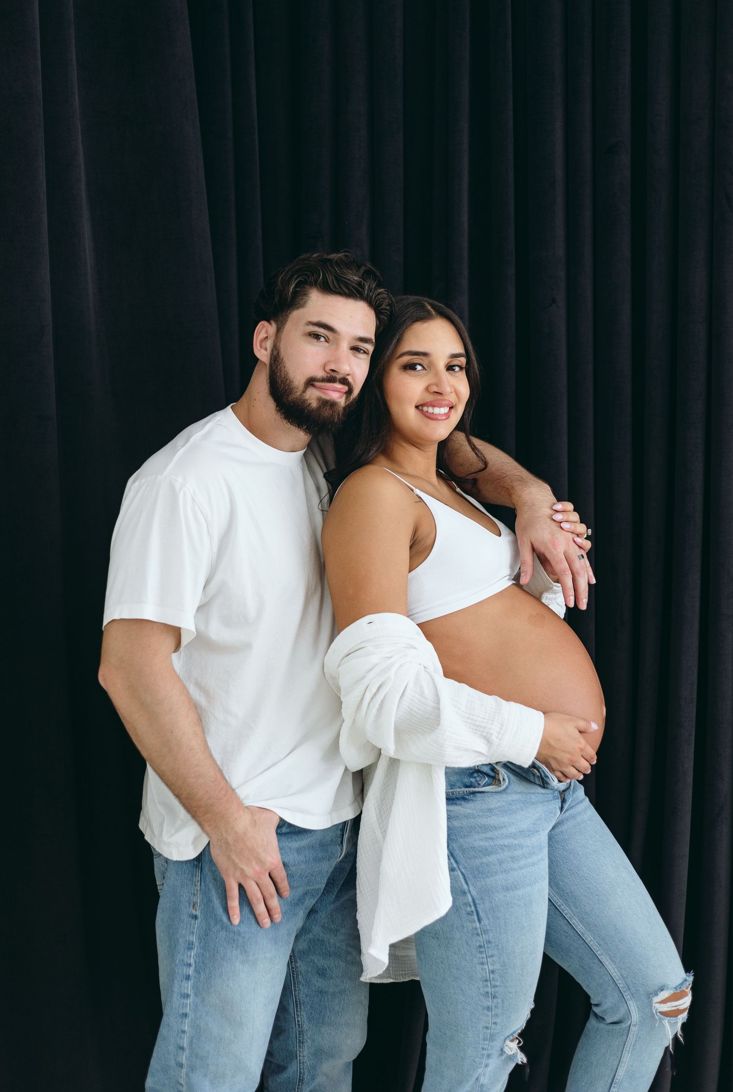 Couple in casual wear posing together against dark backdrop during maternity photoshoot.