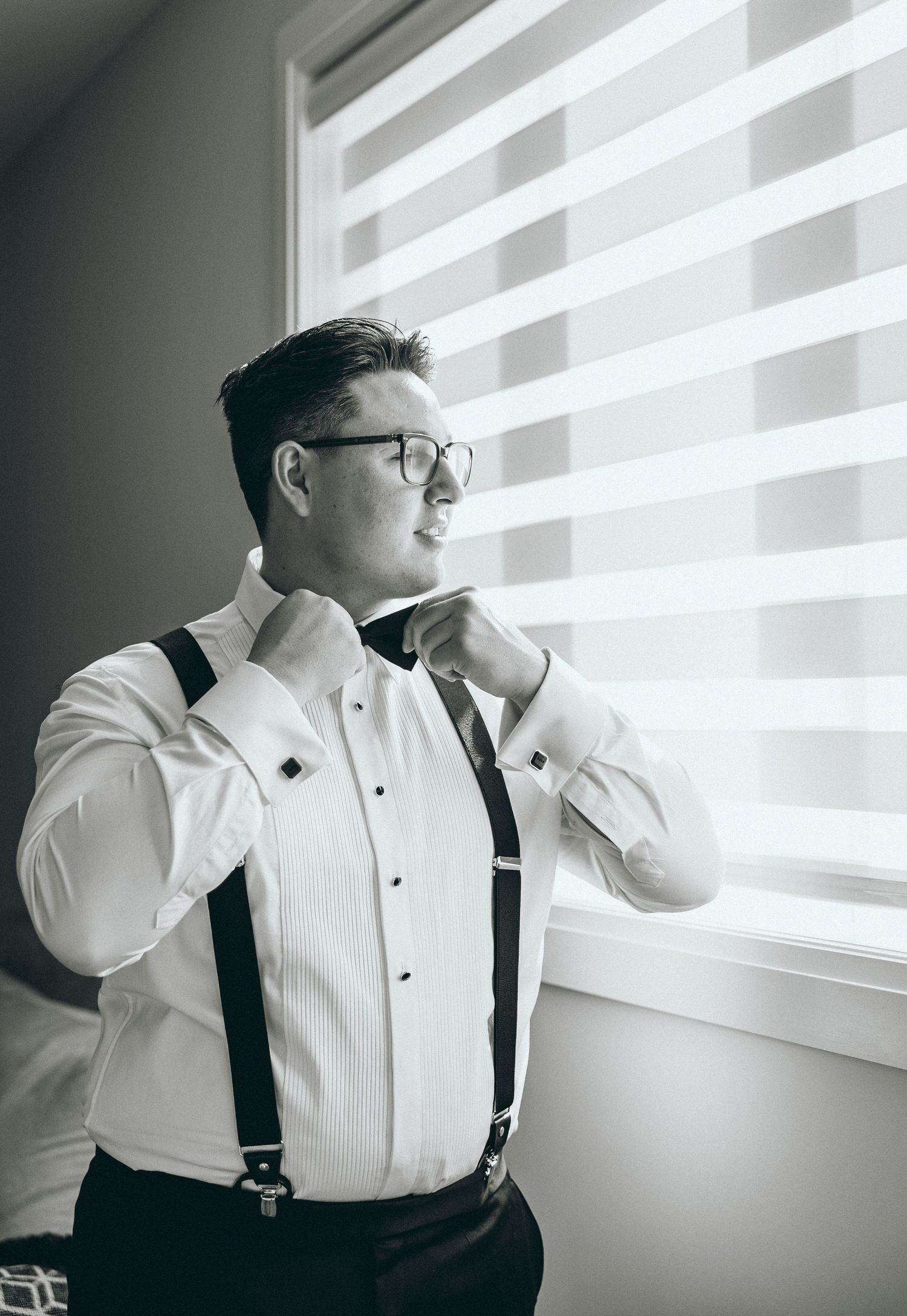 Person in white shirt and suspenders looking out window during wedding preparations.