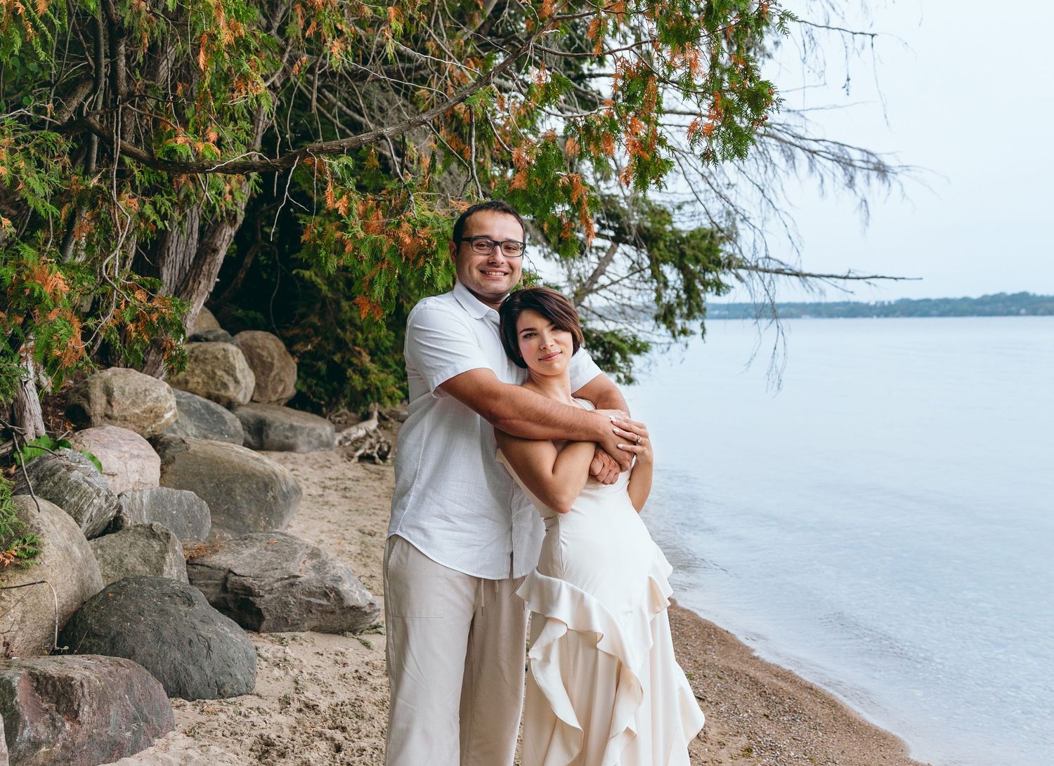 A couple embracing on a rocky beach shore with autumn trees in the background.