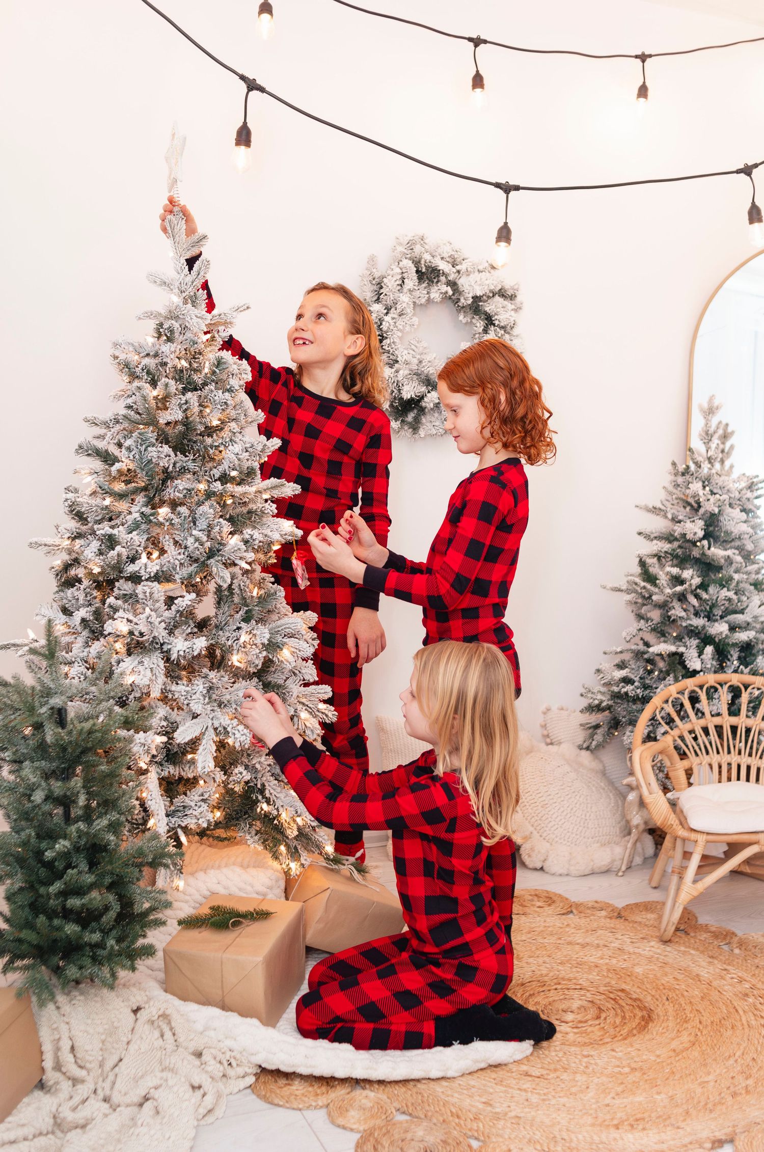 Three girls in matching red plaid pajamas decorating a snow-flocked Christmas tree in a festive studio.