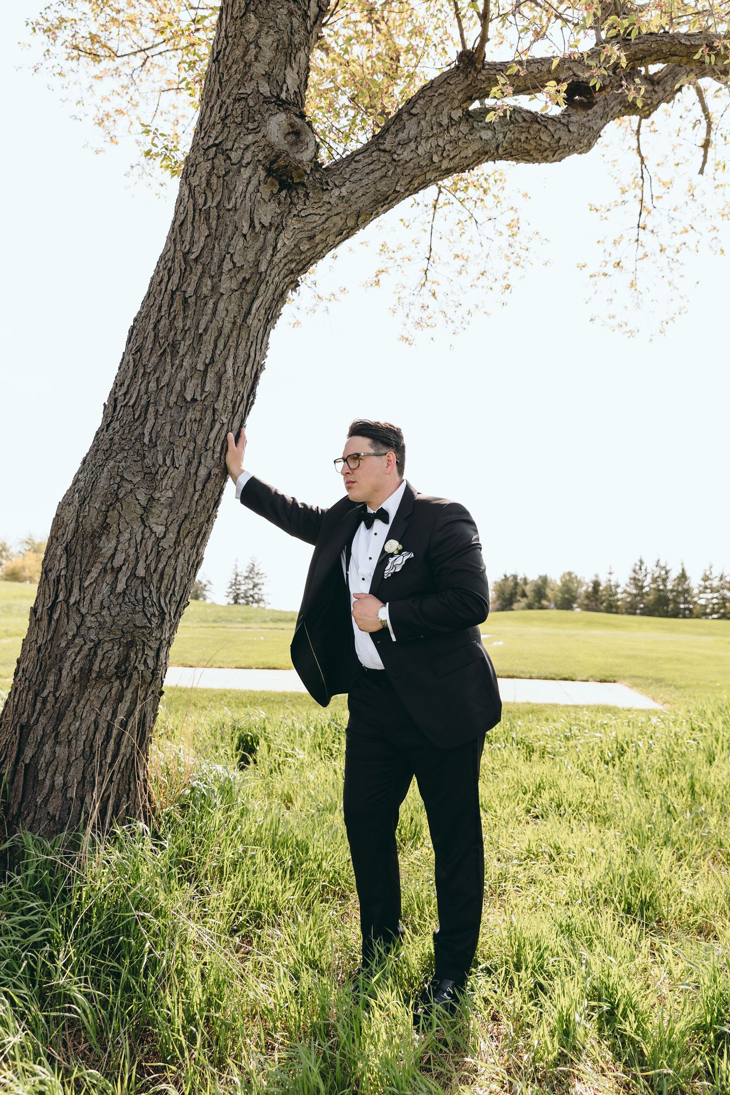 Groom in classic tuxedo poses next to a large tree trunk in an outdoor setting.