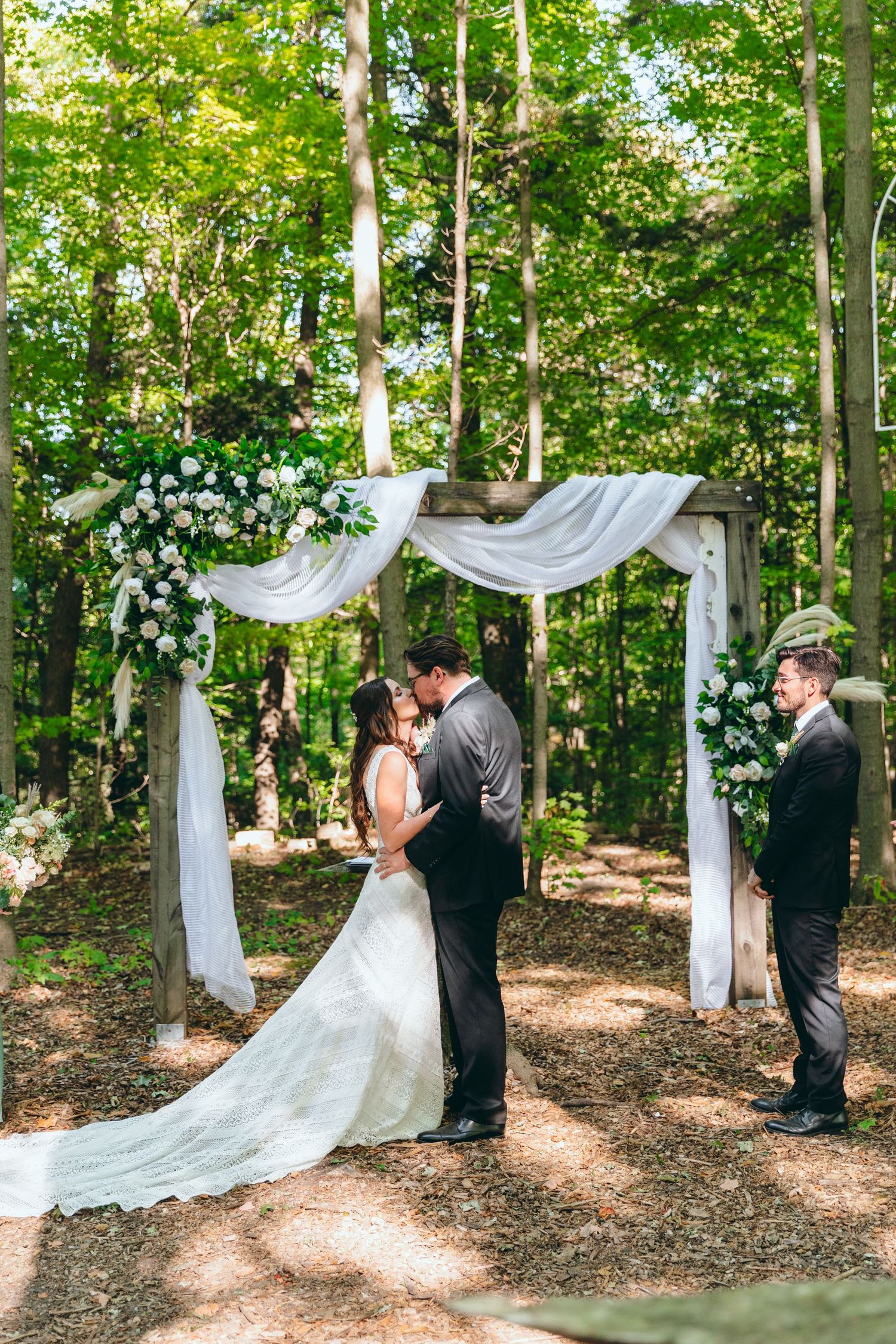 Sunlit forest wedding ceremony with white draped fabric and floral arrangements.