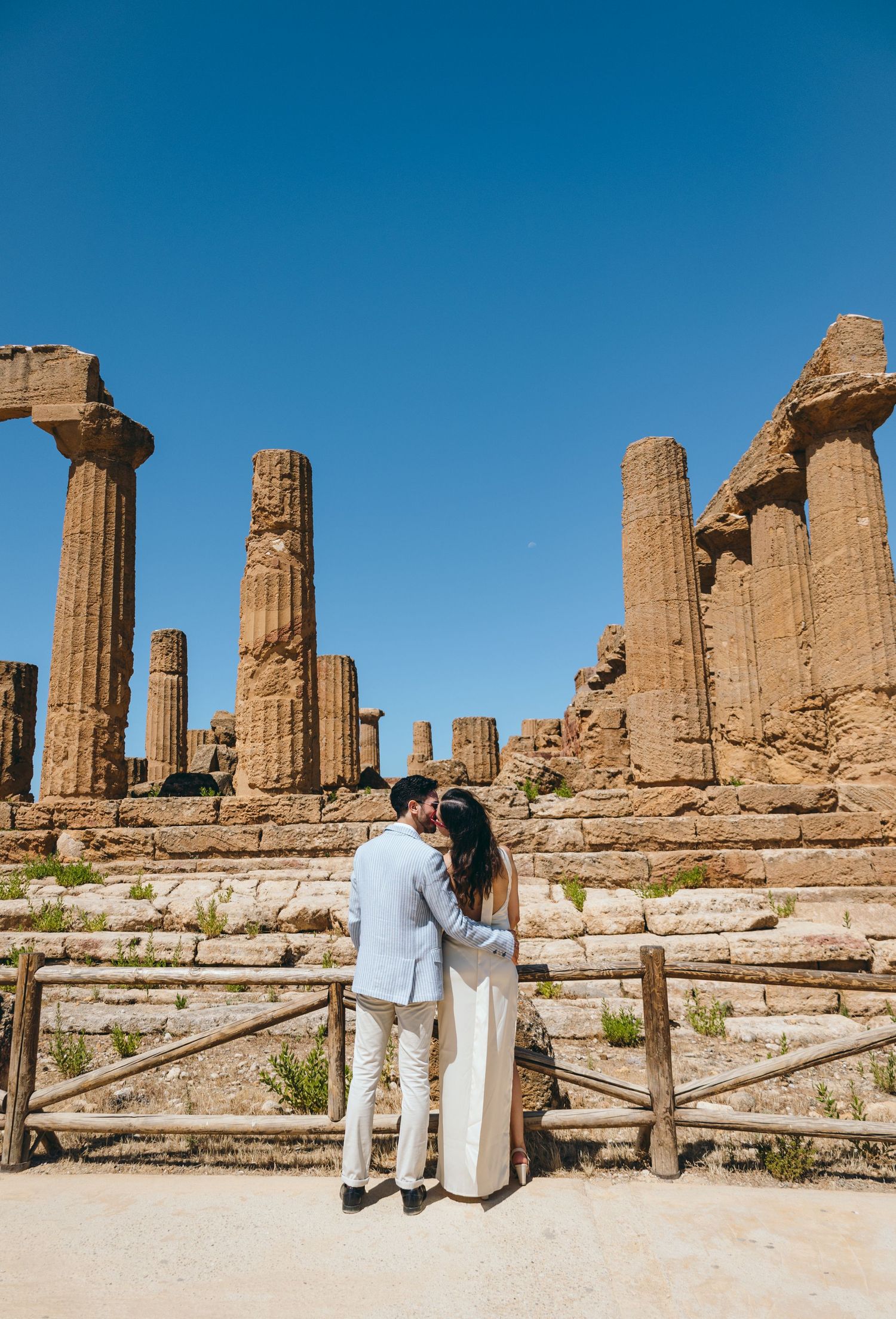 Couple poses at ancient Greek temple ruins with majestic columns under blue sky.