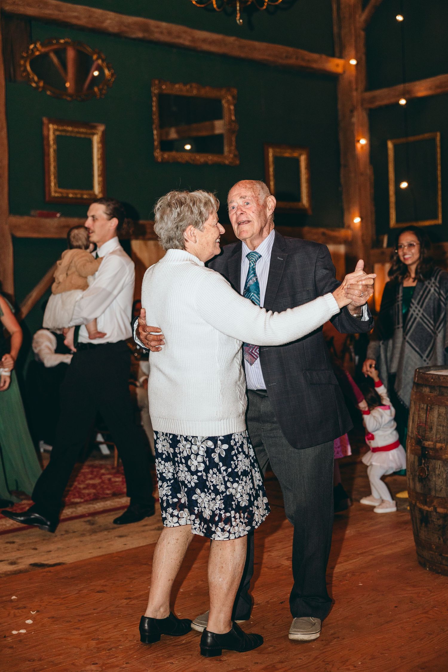 Couples dancing together on wooden floor under chandelier at rustic venue reception.