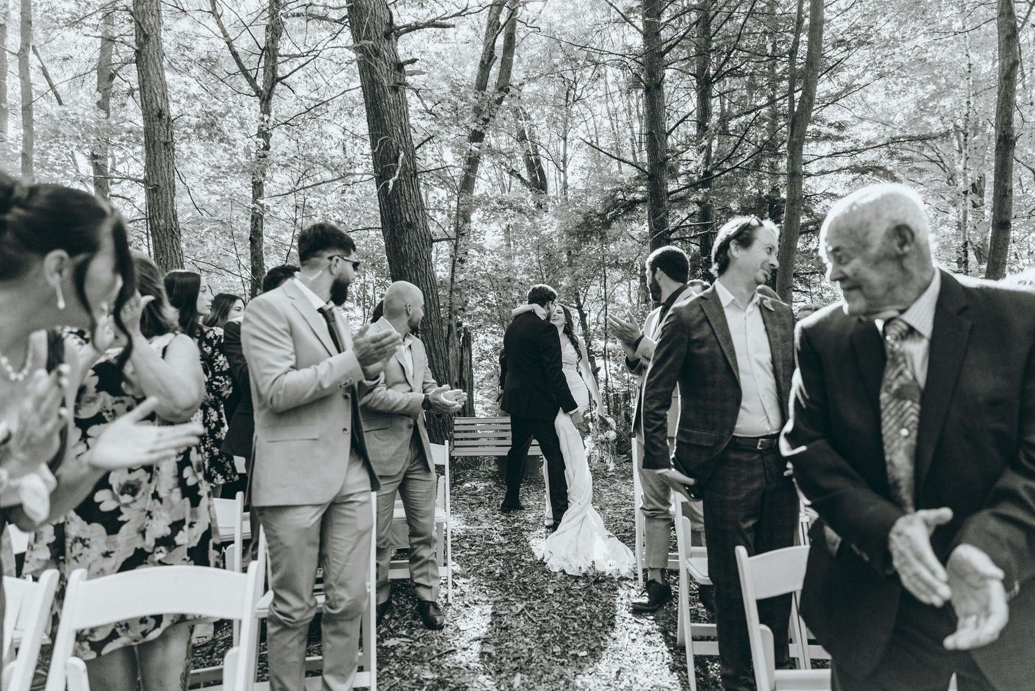Black and white photo of wedding guests standing along an aisle in a wooded outdoor ceremony.