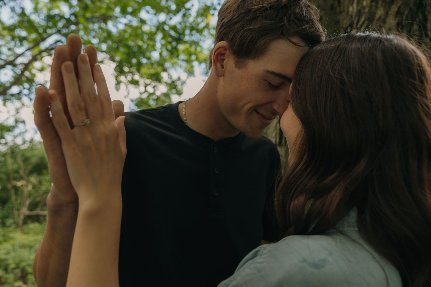 Young couple touching foreheads and holding hands up together near a tree outdoors.