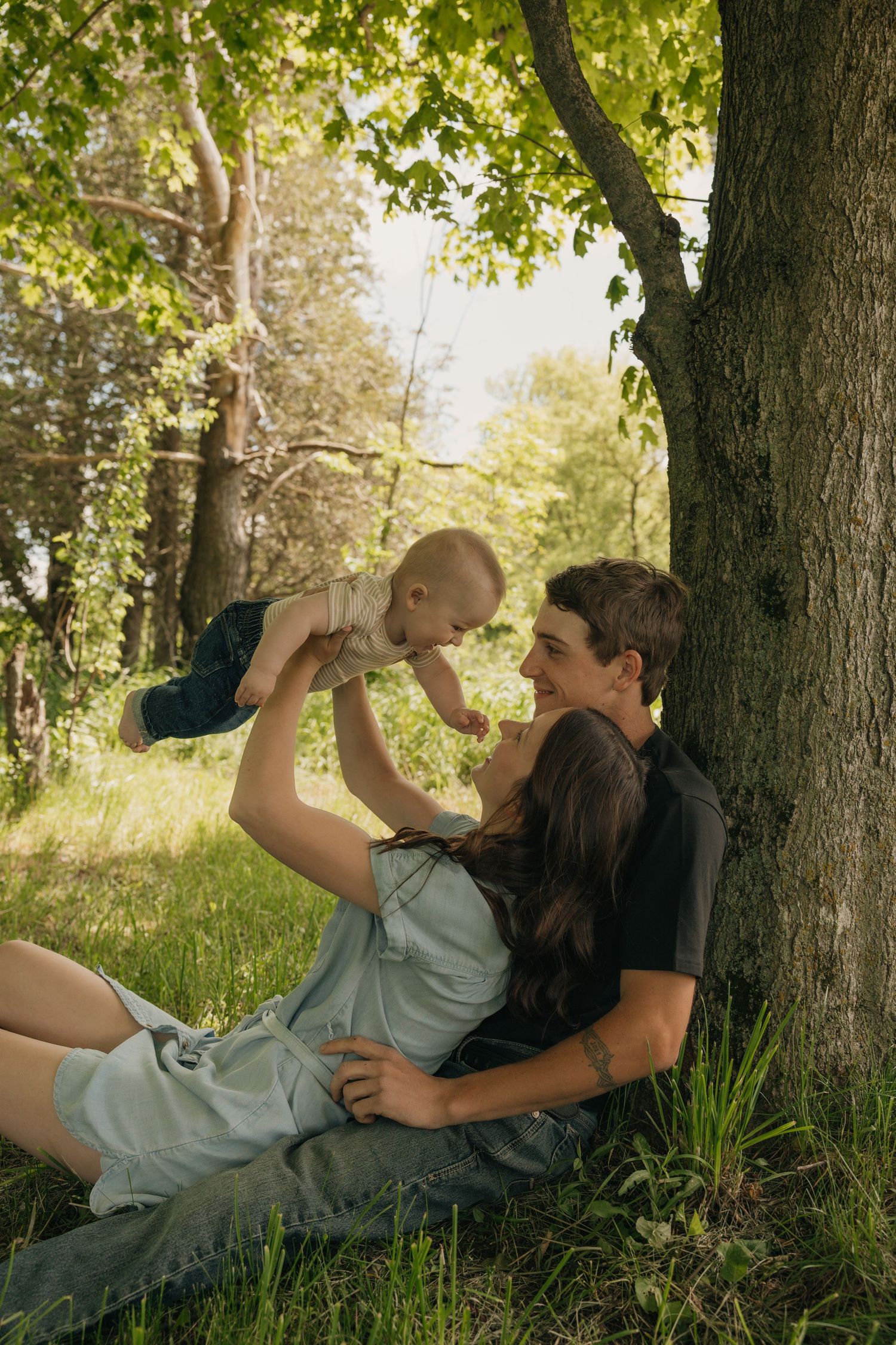 A couple plays with their baby under a large tree in a lush sunlit outdoor setting.