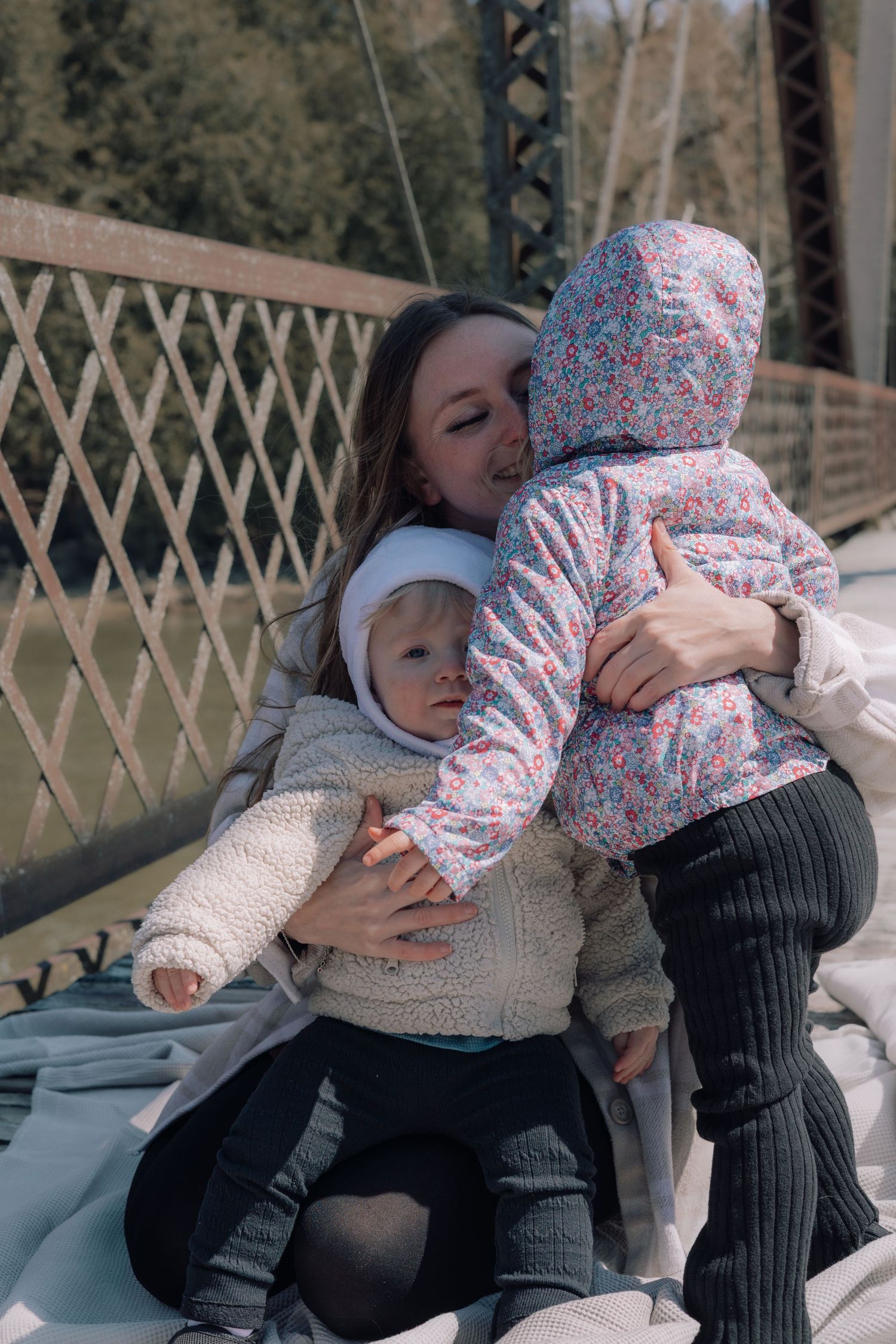 Mother hugging two young children on a blanket on a bridge outdoors.