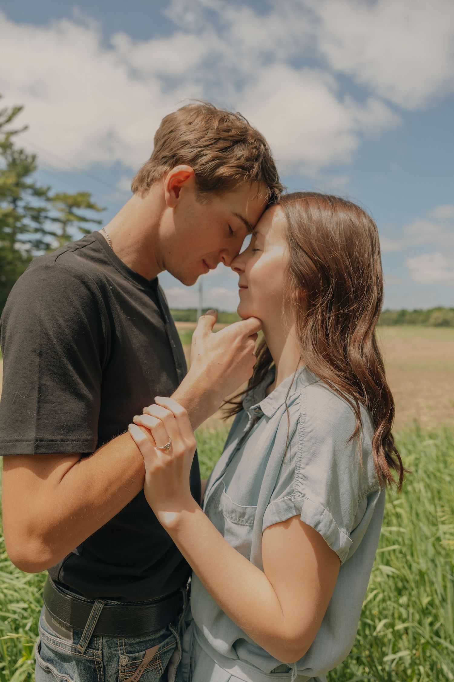 Romantic couple touching foreheads in a green field under a blue sky on a sunny day.