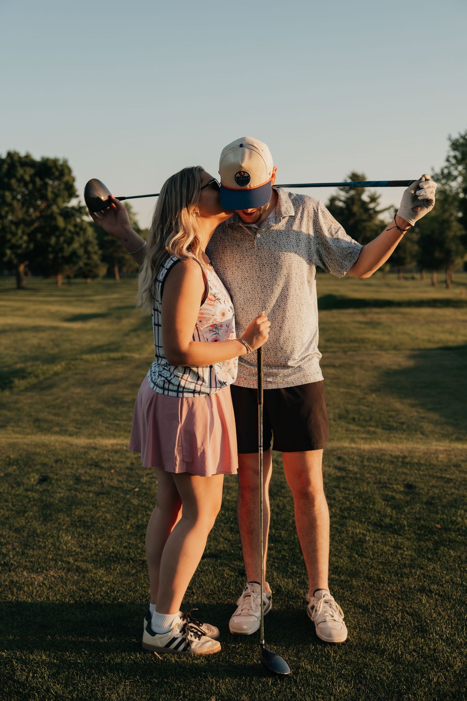Couple sharing a kiss on a golf course at sunset, man holding a golf club across his shoulders.