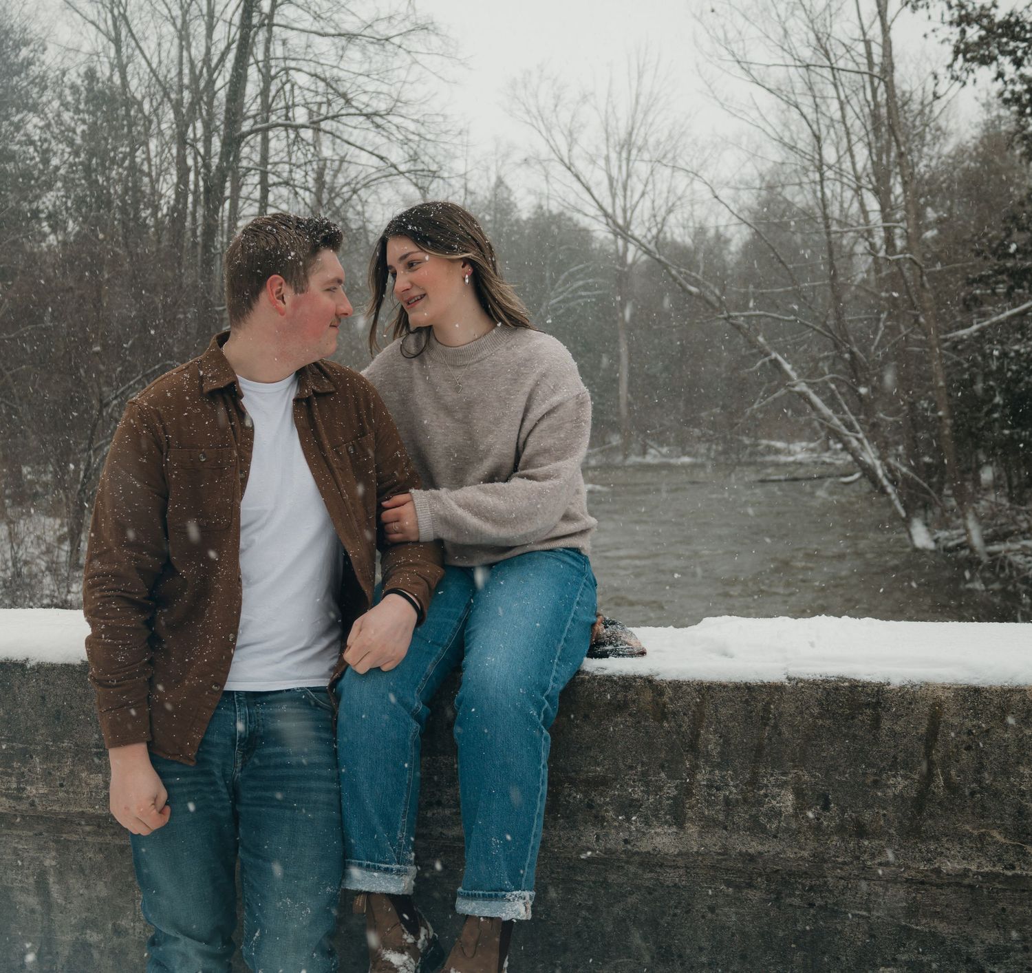 A smiling couple sits together on a snowy stone bridge surrounded by winter trees.