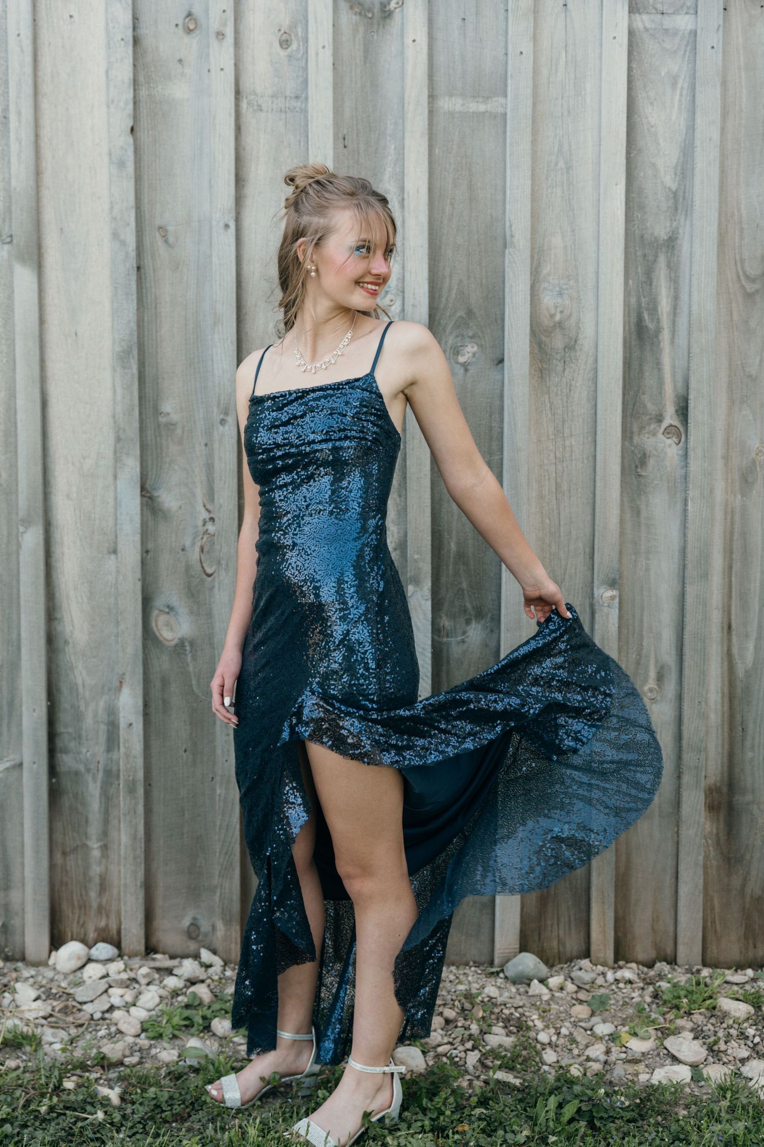 Young woman in stunning navy sequin high-low formal dress posing against rustic wooden fence.