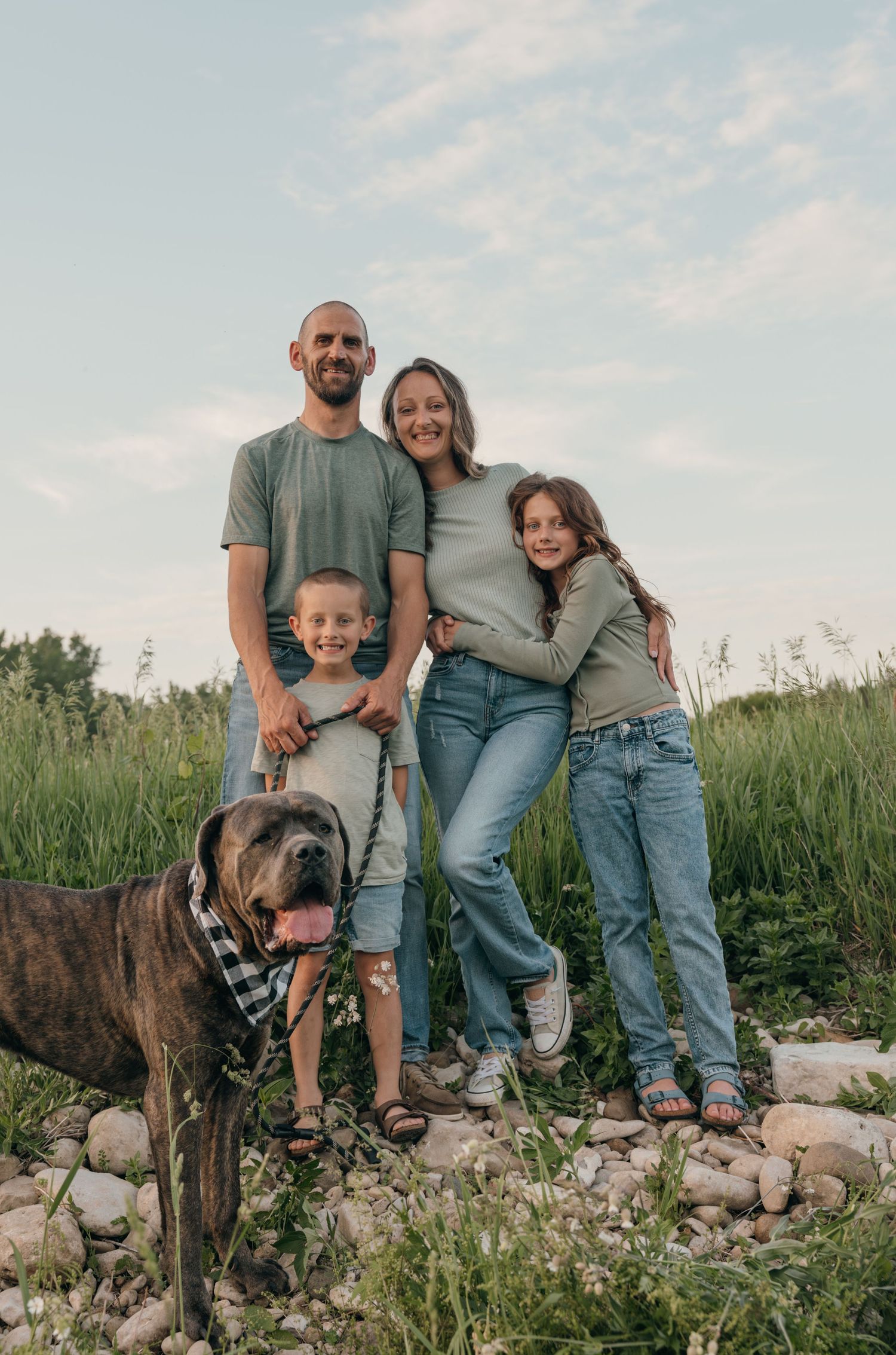 Family of four with a brindle dog in bandana posing on rocky ground in a green meadow.