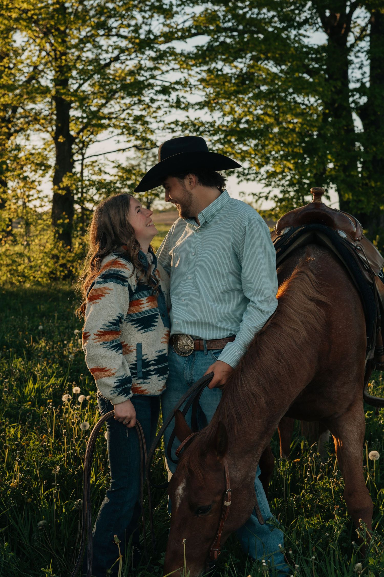 Couple sharing a romantic moment in a sunlit meadow with a brown horse, man wearing cowboy hat and western attire.