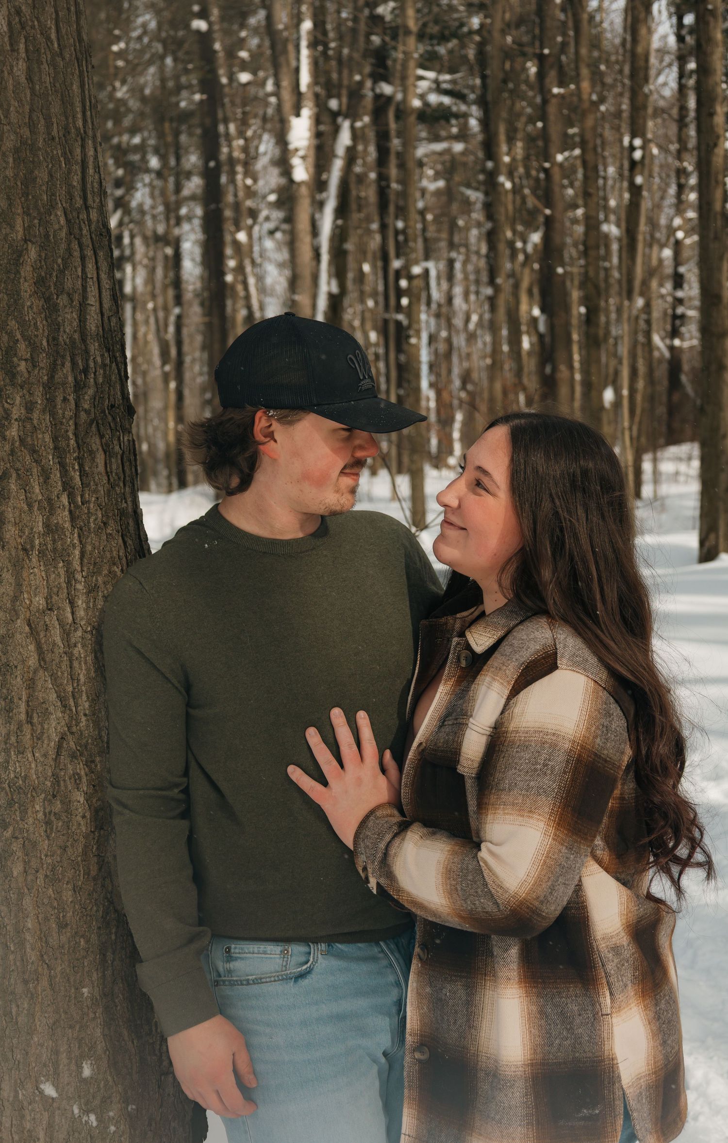 Couple sharing a romantic moment leaning against a tree in a snowy forest.