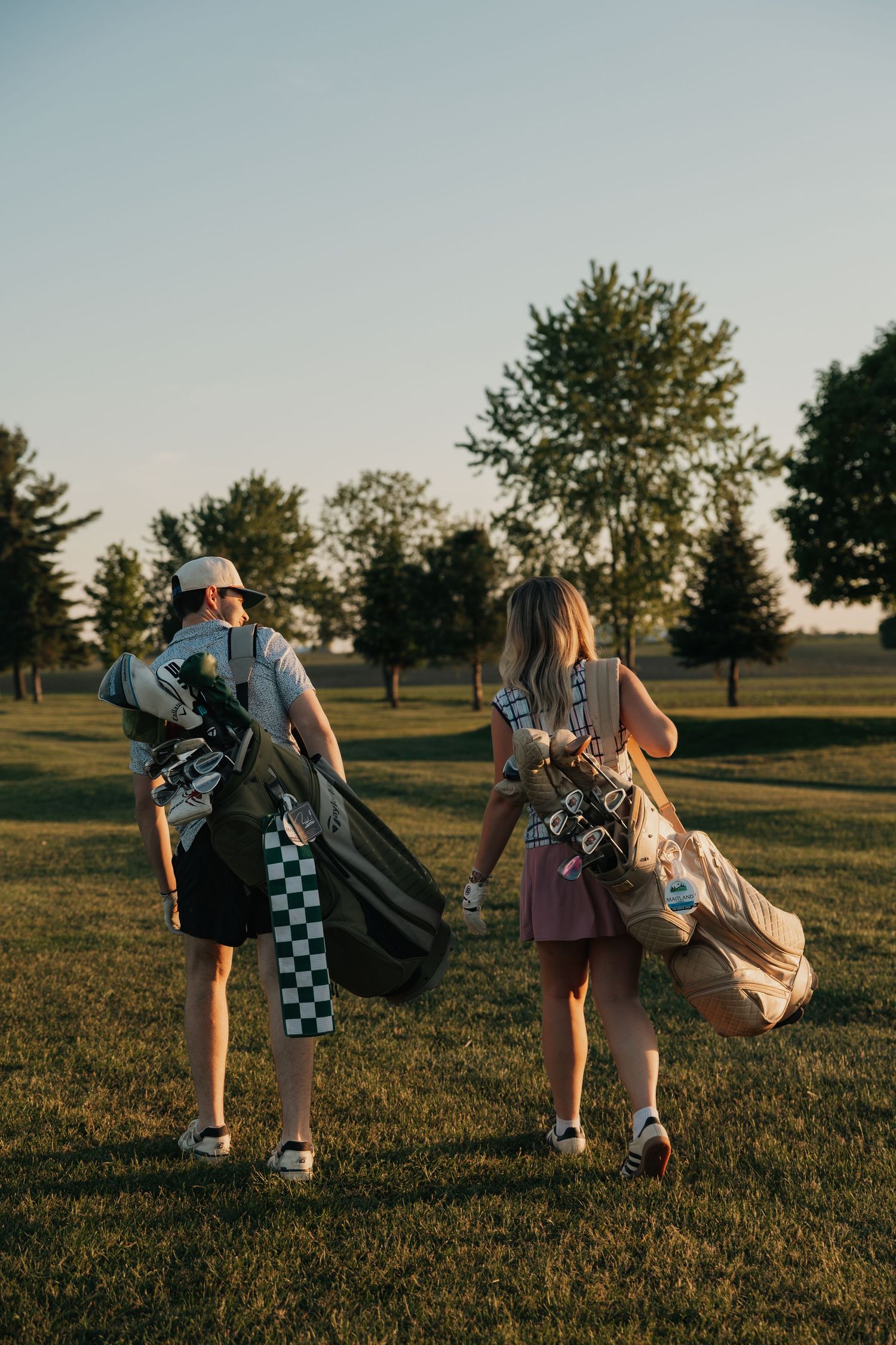 Two golfers carrying golf bags walk across green fairway at golden hour.