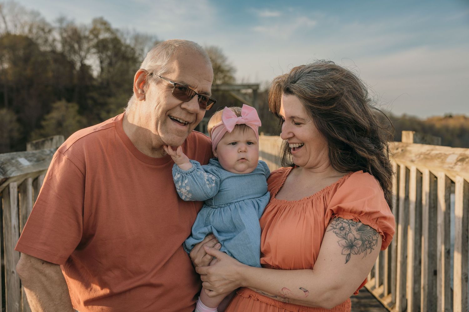 Grandparents smiling and holding a baby girl with a pink bow outdoors.