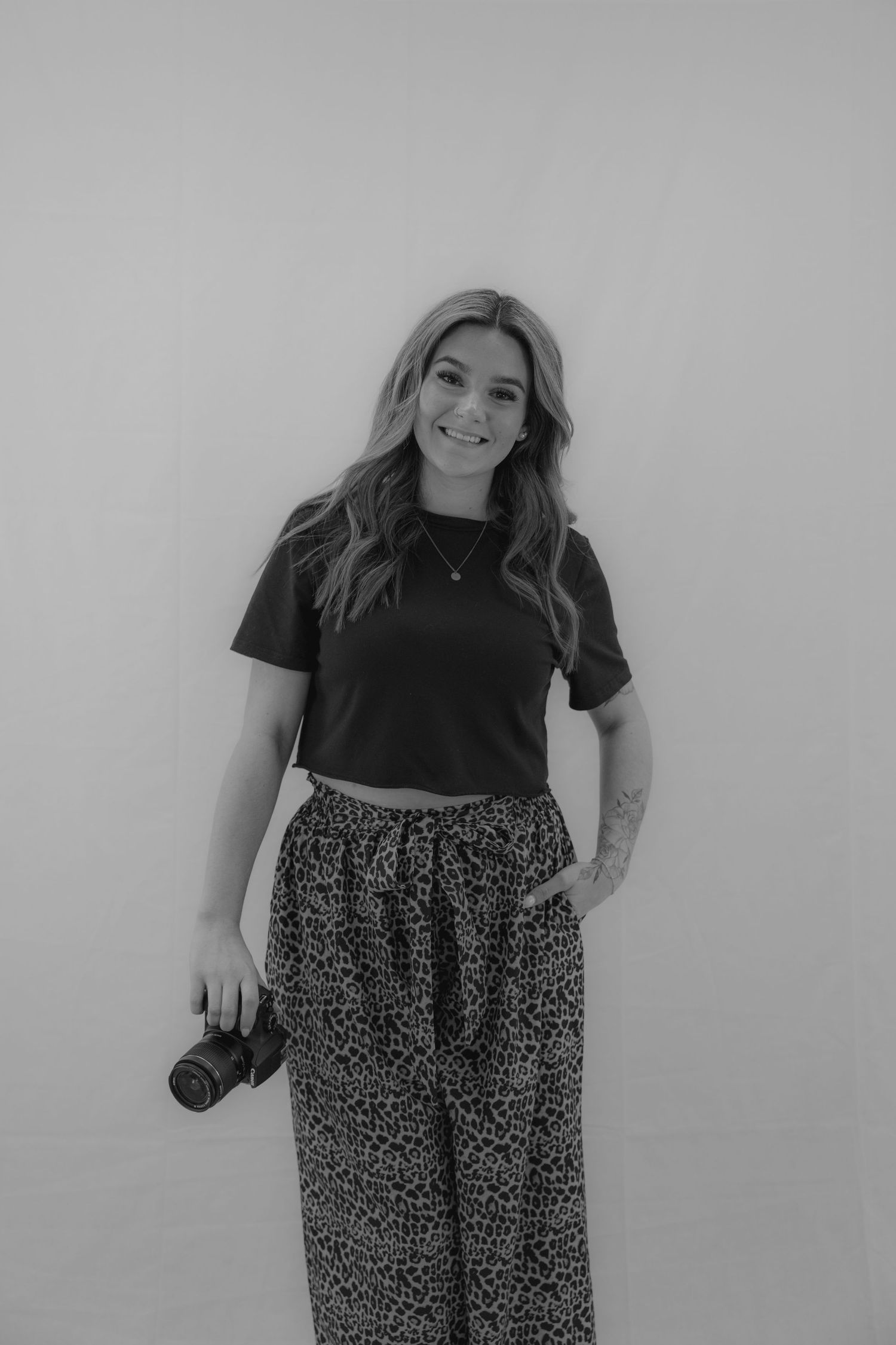 A smiling woman in a black crop top and leopard print pants holds a camera in a black and white portrait photo.