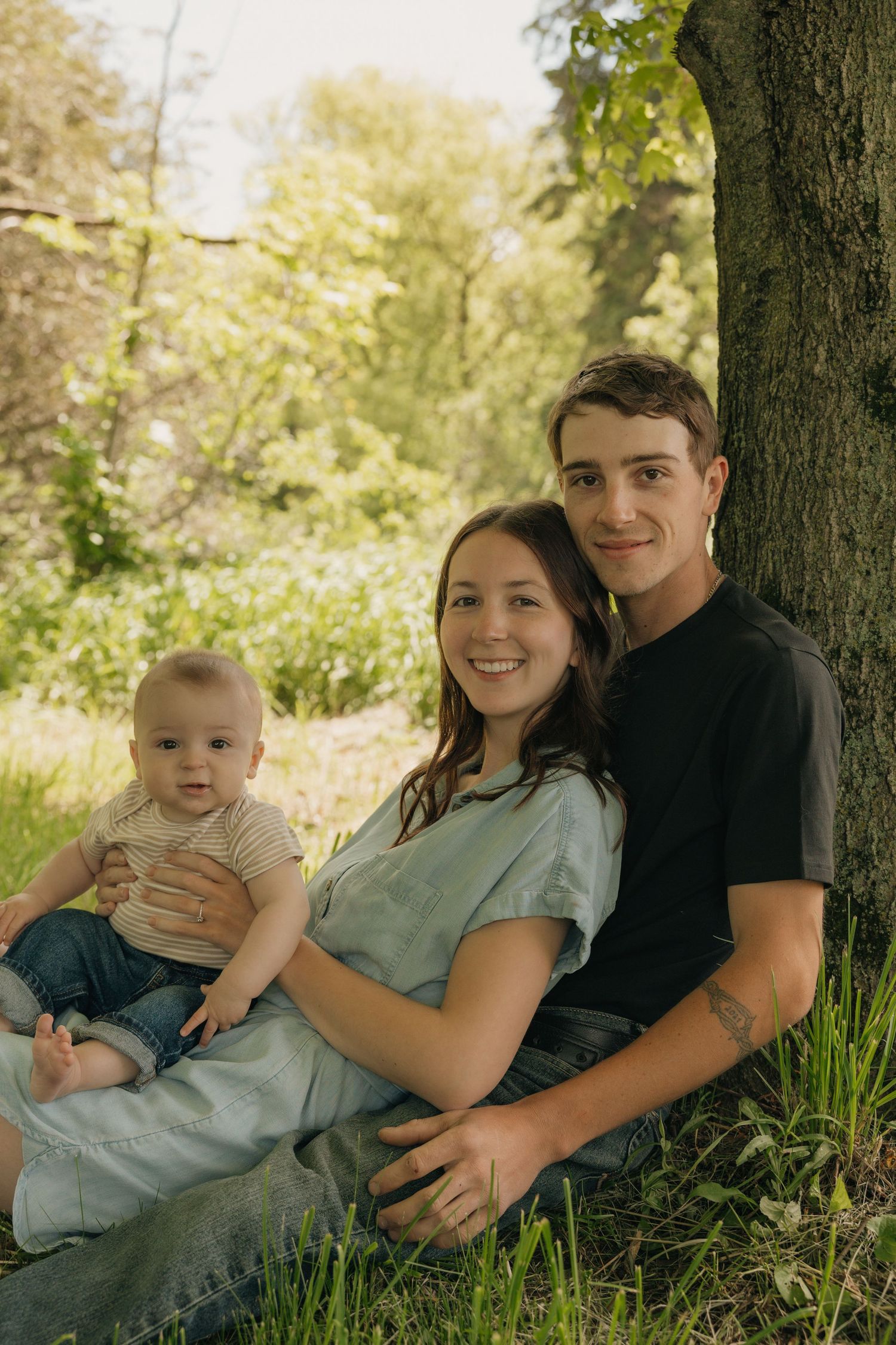 A young family with a baby sits together against a tree in a sunny green park.