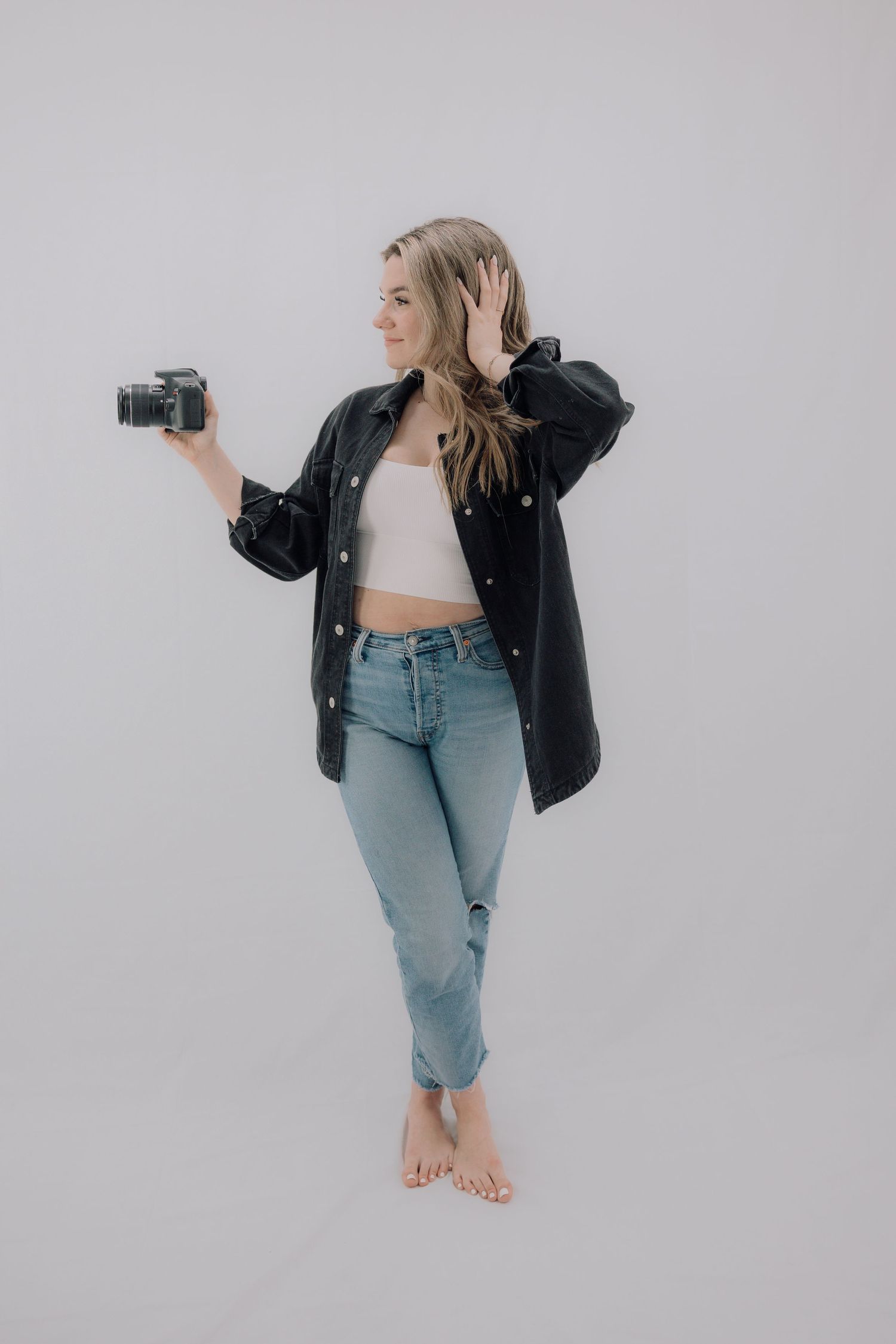 Blonde woman in black jacket and jeans holding a camera while posing against a gray background.
