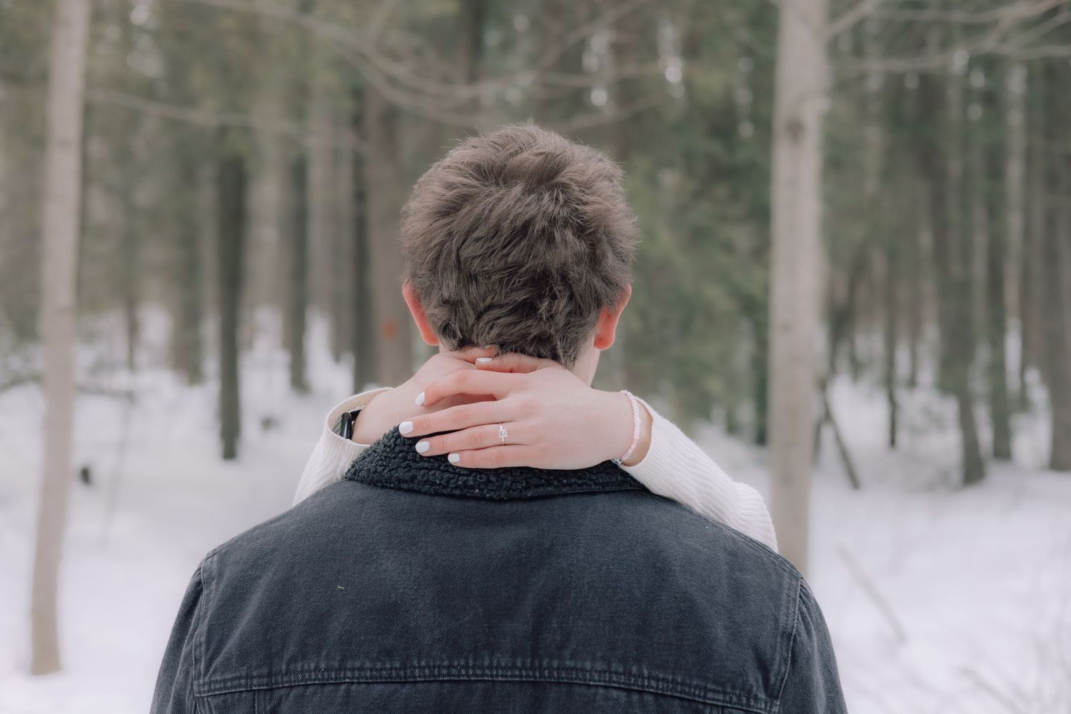 A woman rests her hand on a mans neck from behind in a snowy winter forest scene.