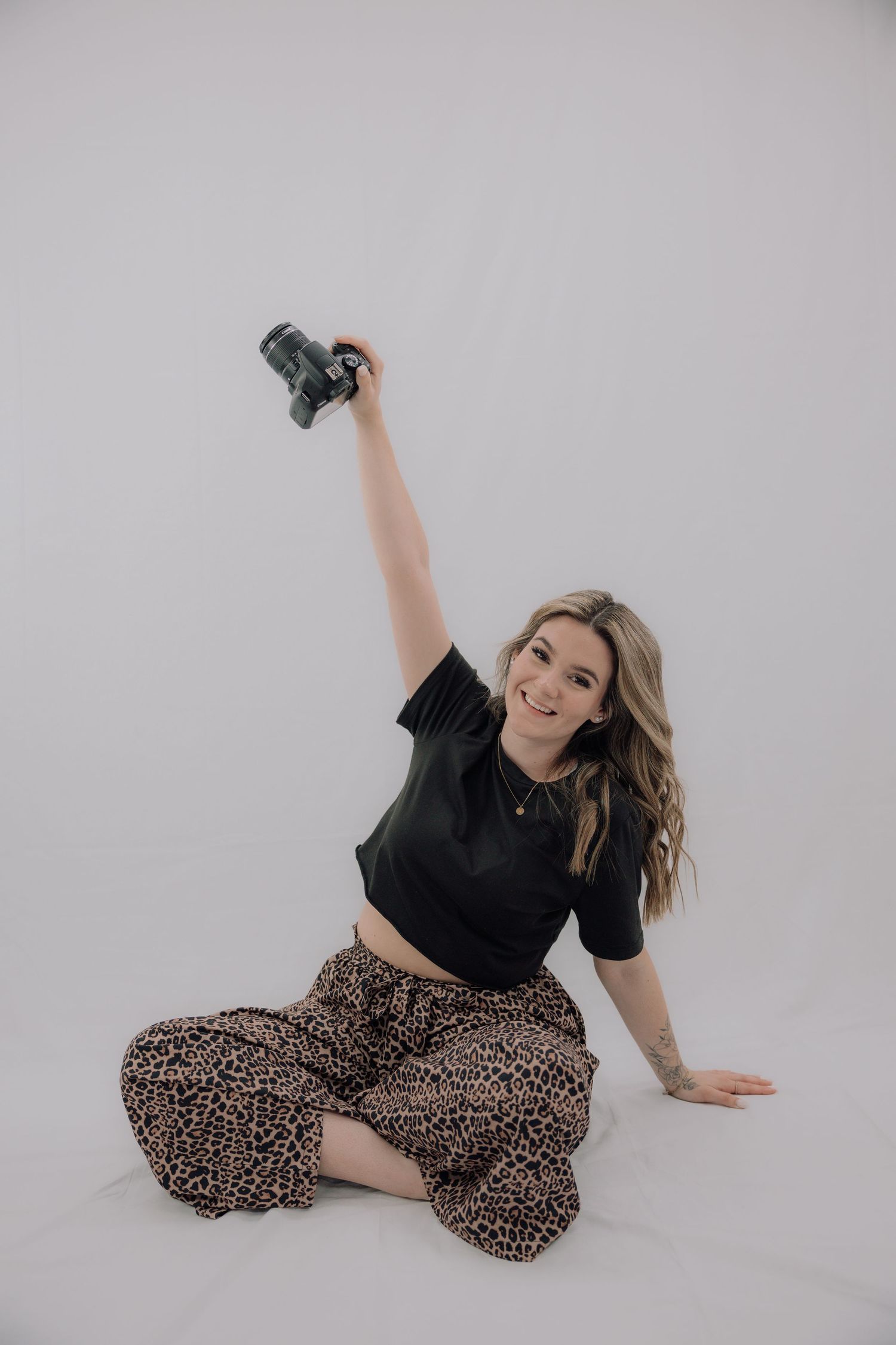 A smiling woman in a black crop top and leopard print pants sits on the floor holding a camera above her head.