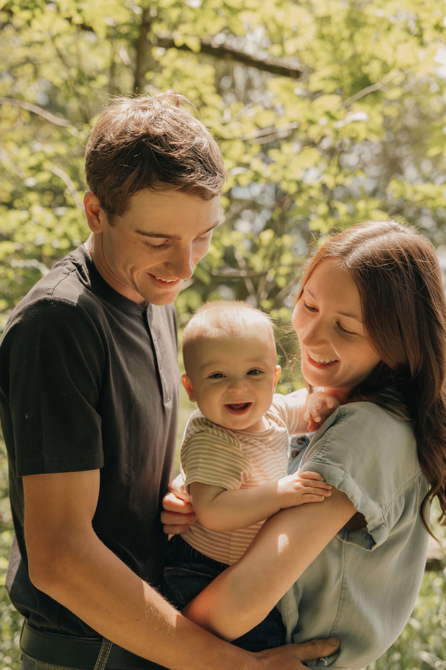 Young couple smiling and holding their laughing baby outdoors in a sunny green park setting.