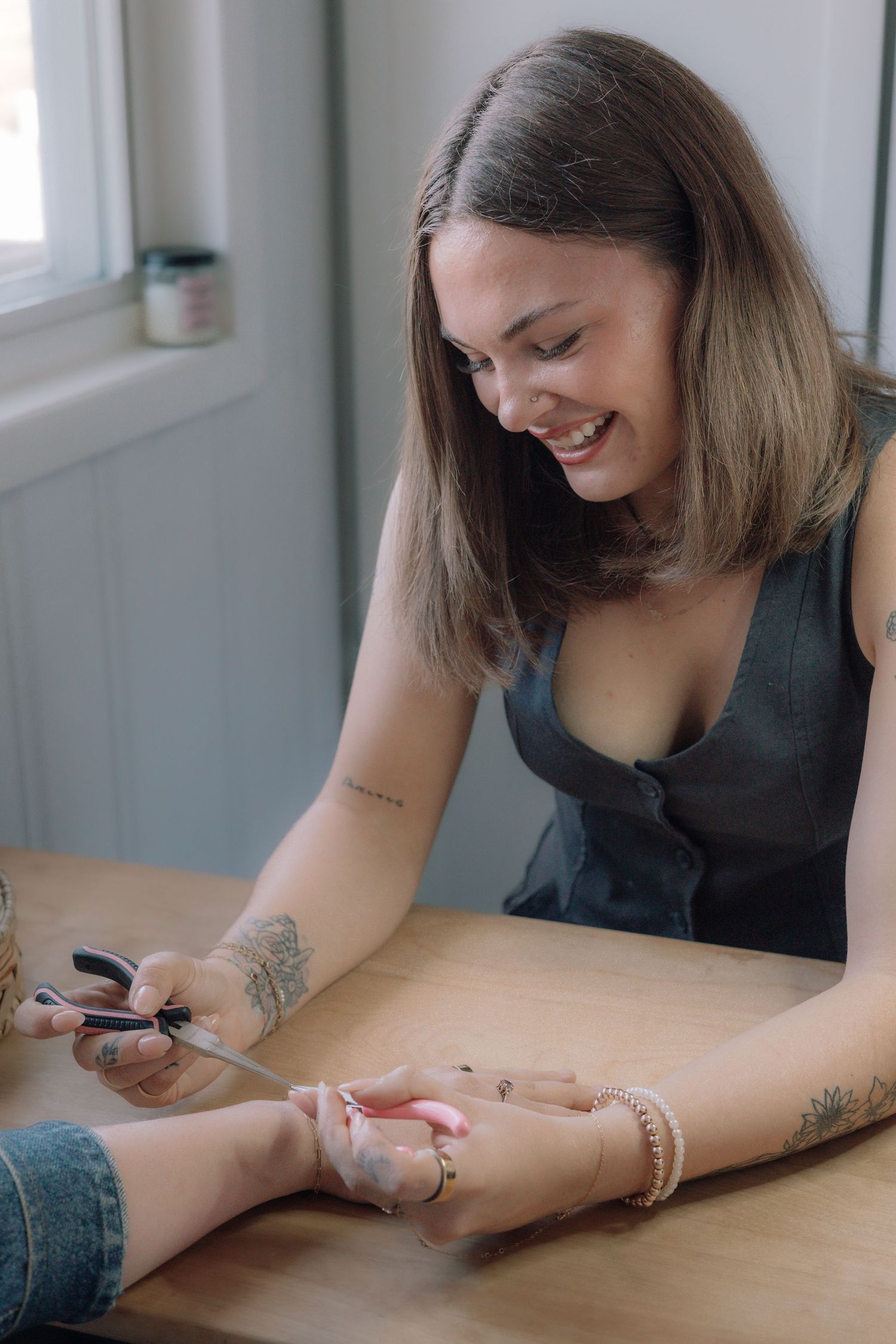 Smiling tattooed woman performing nail care service on client at wooden table in bright salon setting.