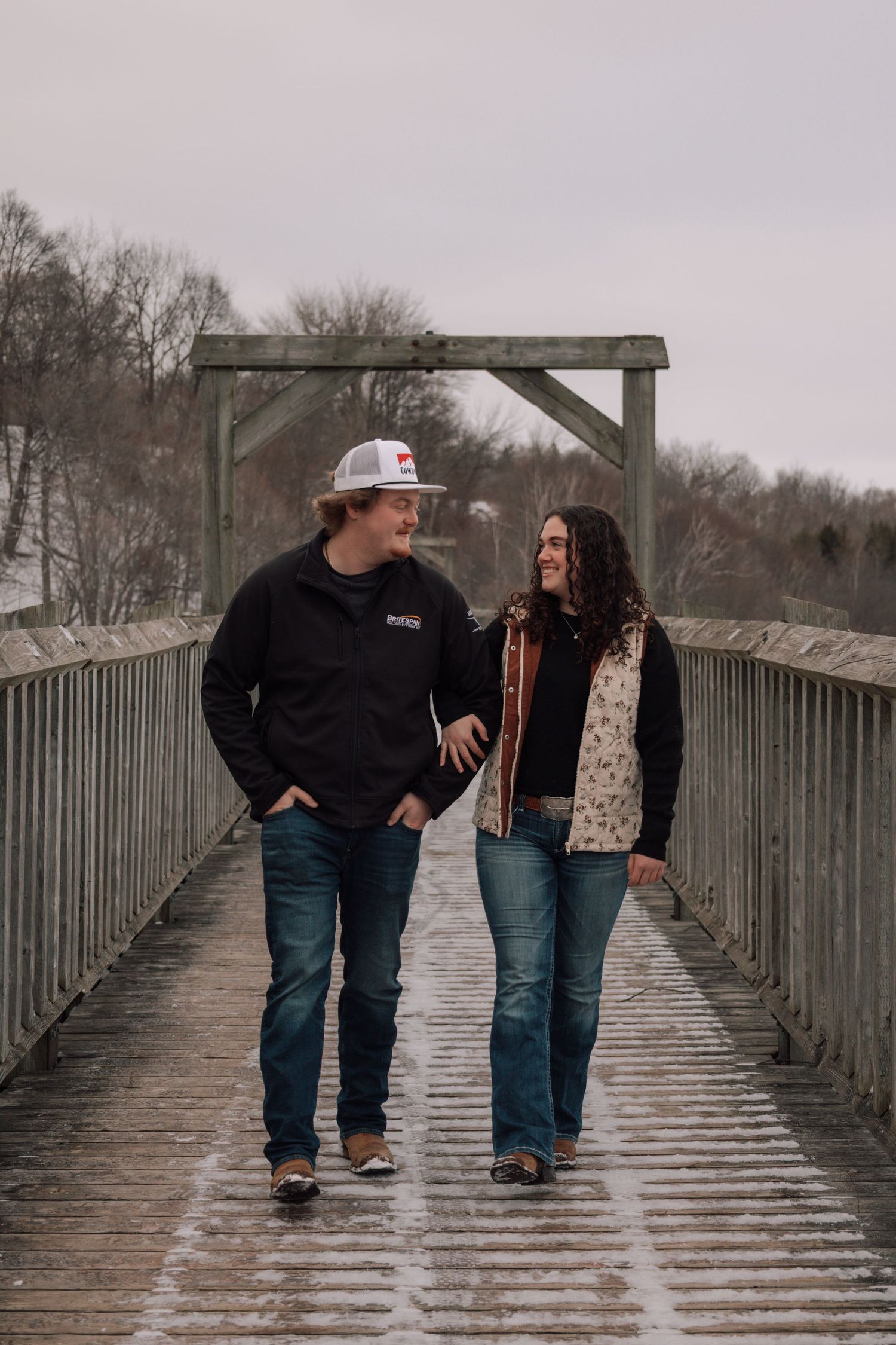 Couple walking and smiling at each other on a snow-dusted wooden bridge.