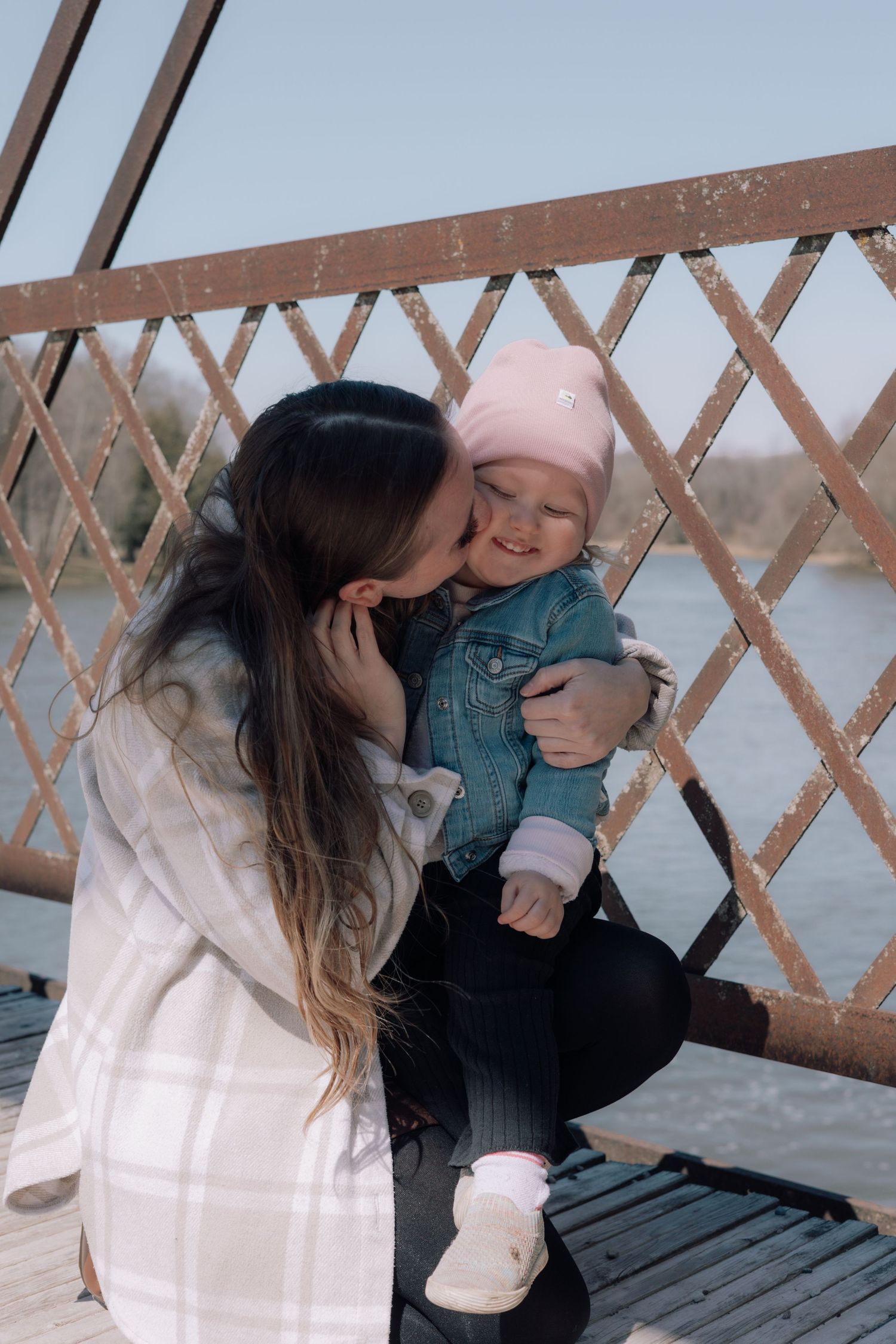 Mother kissing her smiling baby wearing a pink hat on a bridge by water.